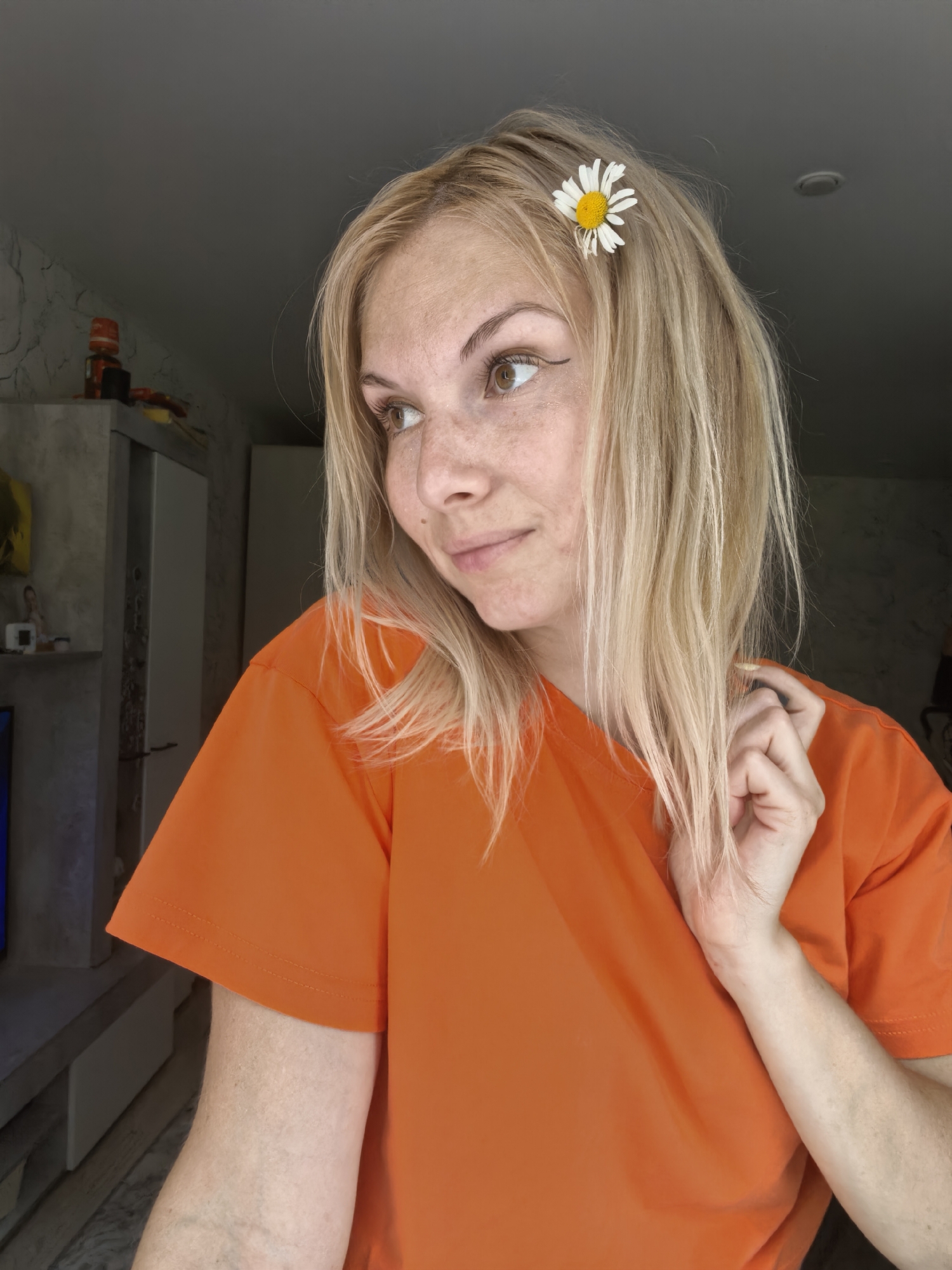 A woman with blonde hair and freckles, adorned with a daisy in her hair, is captured in a close-up shot. She is wearing an orange t-shirt and looking to her left with a slight smile. Her right hand is raised, with fingers touching her hair near her cheek. The background appears to be an indoor setting, possibly a room in a residential building in Sterlitamak, Russia, suggested by the presence of furniture and a wall with textured wallpaper. The lighting is natural, coming from a window or doorway on the left side of the frame, casting a soft glow on the woman's face.
