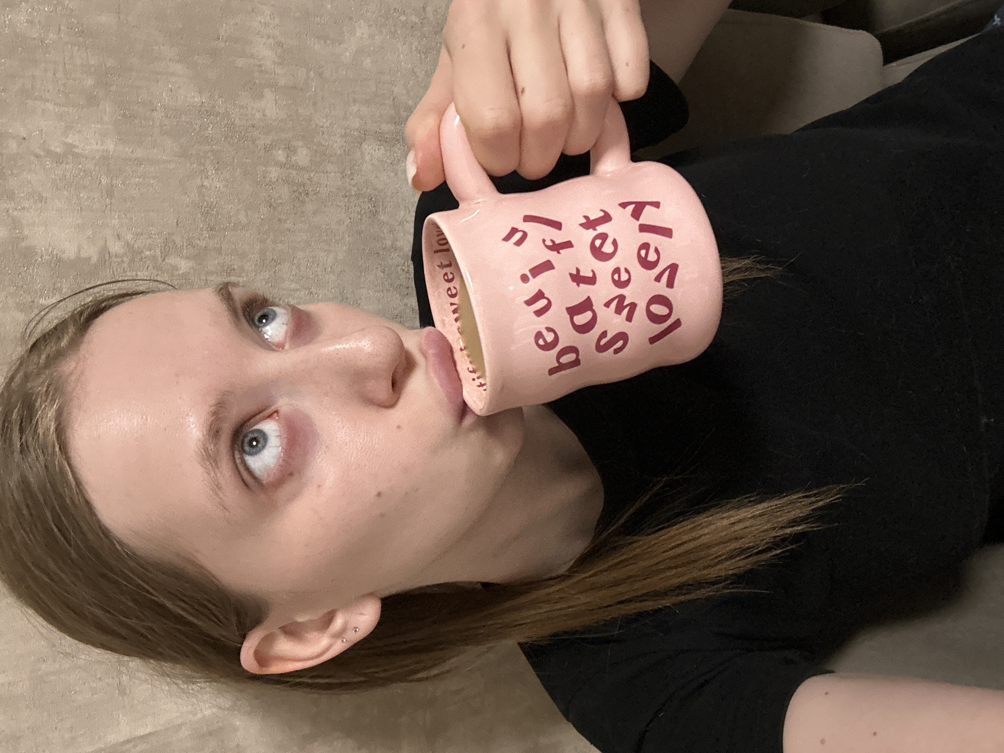 A young woman with fair skin and light brown hair is lying on a beige surface, possibly a couch or bed. She is wearing a black shirt. Her head is tilted back, and she is looking up and to the left. She is holding a pink mug with the words "lovely," "sweet," and "beautiful" printed on it. The mug is touching her lips as if she is about to drink from it. The scene provides no information to infer the location of Chaikovsky, Russia.