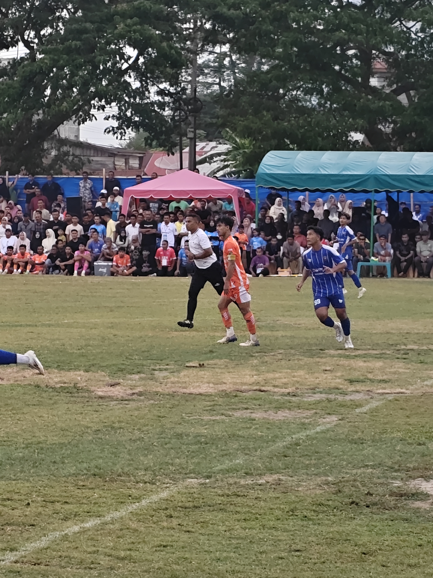 This media file captures a football match in progress at Desa Meunasah Blang, Indonesia. The scene is set on a grassy field with white boundary lines, indicating a formal playing area. In the foreground, three male football players are the central subjects. One player in a white shirt and black shorts is in mid-stride, seemingly focused on the game. Another player, wearing an orange and white striped jersey and shorts, is also in motion. The third player, in a blue and white striped jersey with the number 99, runs towards the right side of the frame, his expression suggesting concentration.
Behind the players, a large crowd of spectators is gathered, seated or standing behind a barrier. They are watching the match intently. The spectators are a mix of men, women, and children, many wearing casual clothing. Some are sheltered by brightly colored tents, a pink one on the left and a teal one on the right, which may serve as team shelters or spectator areas. The background reveals lush green trees and some buildings, suggesting a semi-urban or rural setting. The sky appears overcast, hinting at a cloudy day. The overall atmosphere seems to be one of community engagement and sporting enthusiasm, typical of a local football event. No specific text is clearly legible on the players' uniforms or the surroundings, though small logos or text might be present on the players' kits, too small to discern. The time of day appears to be daytime, possibly late afternoon, given the lighting conditions.