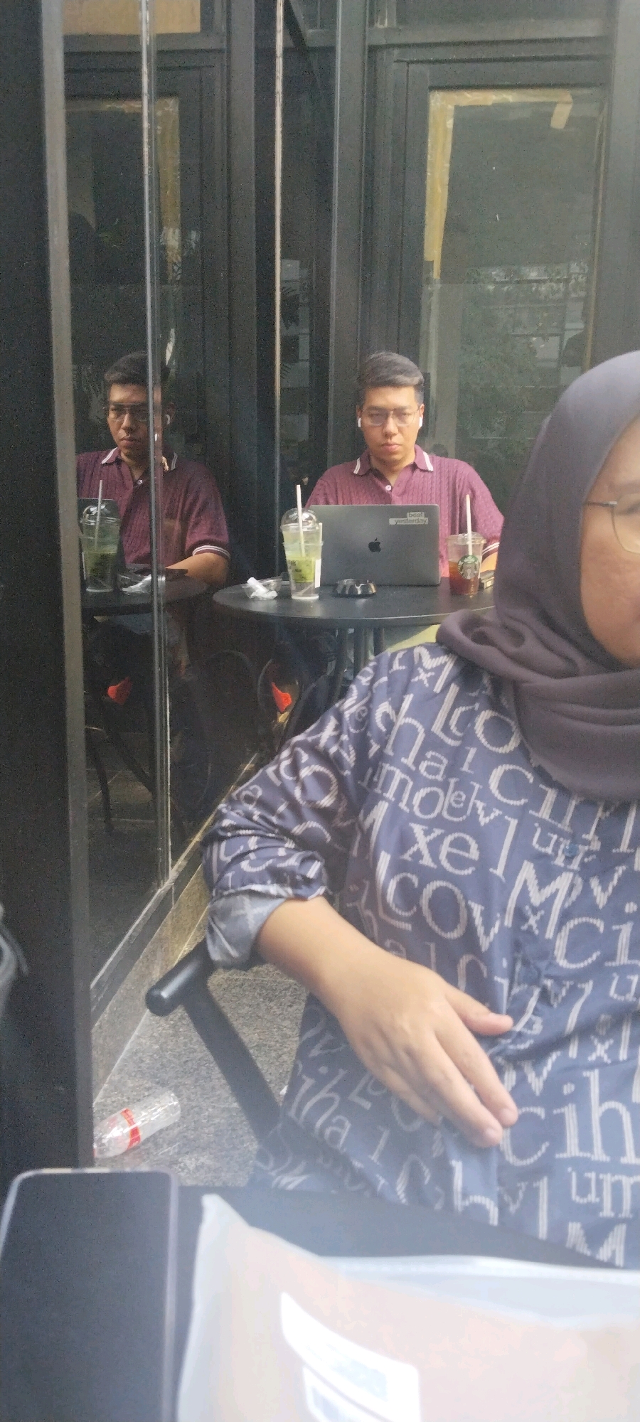The image shows a man sitting at a table with a laptop and drinks, reflected in glass. He is wearing a maroon polo shirt and glasses, with wireless earbuds in his ears. Next to him on the table are two drinks, one in a clear plastic cup with a straw, and another in a cup with a Starbucks logo. A partially visible woman wearing a hijab and a patterned shirt is in the foreground. A plastic water bottle is on the ground near the table. The scene appears to be an outdoor seating area of a cafe, possibly in South Jakarta, Indonesia, given the context.