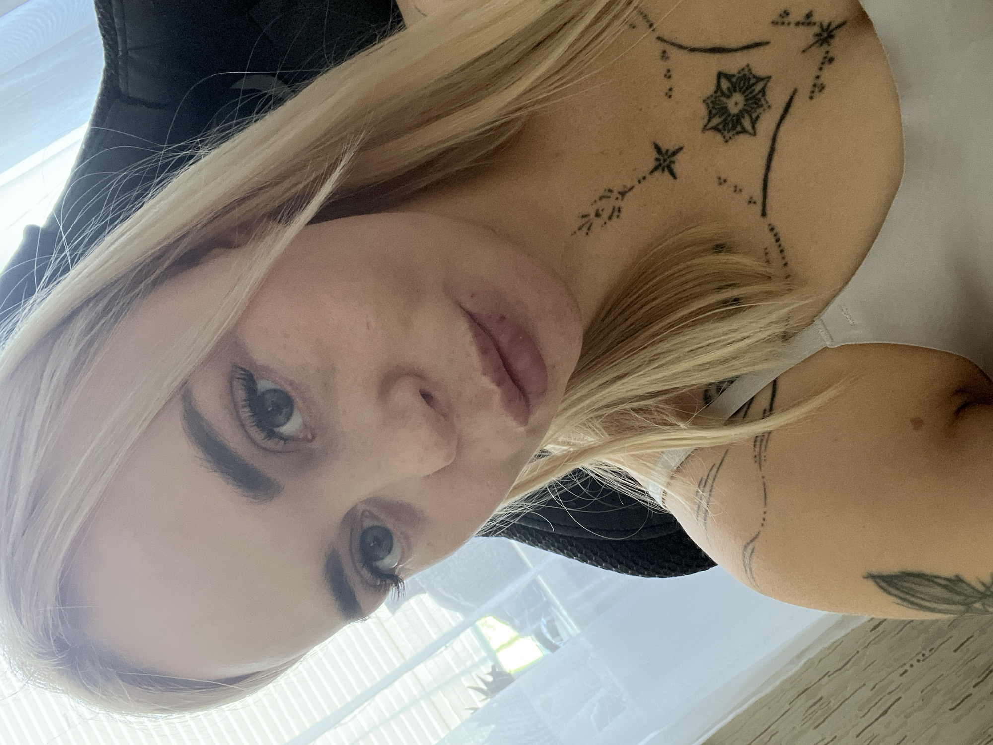 A close-up, waist-up selfie shows a woman with blonde hair and blue eyes. She is wearing a light-colored tank top. She has tattoos on her chest and left shoulder. The background appears to be indoors, with blinds visible on a window to the left, and a dark chair behind her. The lighting is bright, suggesting daytime.