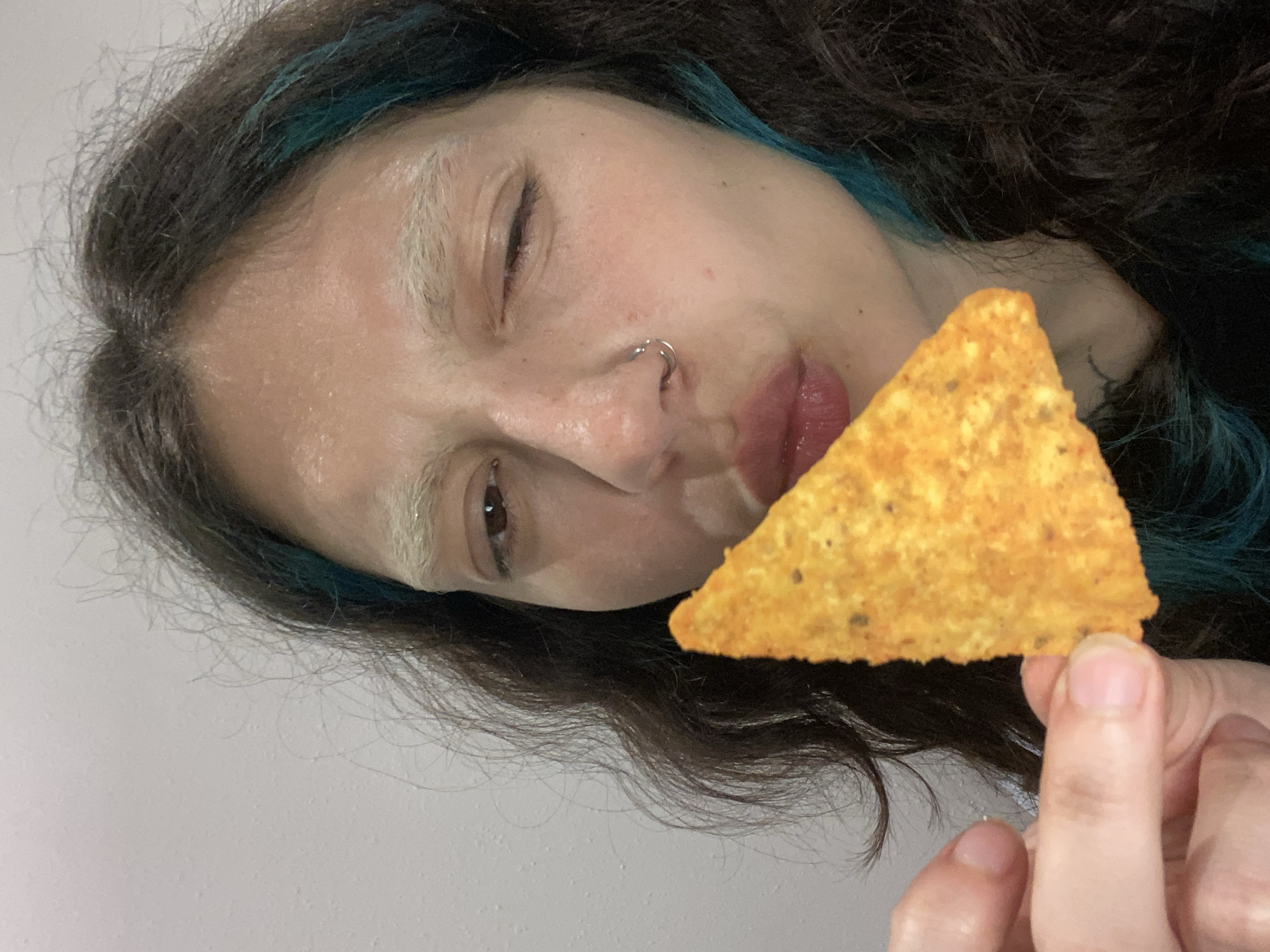 A person with dark brown hair highlighted with blue and bleached eyebrows is holding a triangular tortilla chip up to the camera, partially obscuring their face. The person is winking. They have a silver hoop nose ring in their left nostril. Their fingernails are short and unpainted. The background is a plain white wall. There is no indication the image was taken in Gijón/Xixón, Spain.