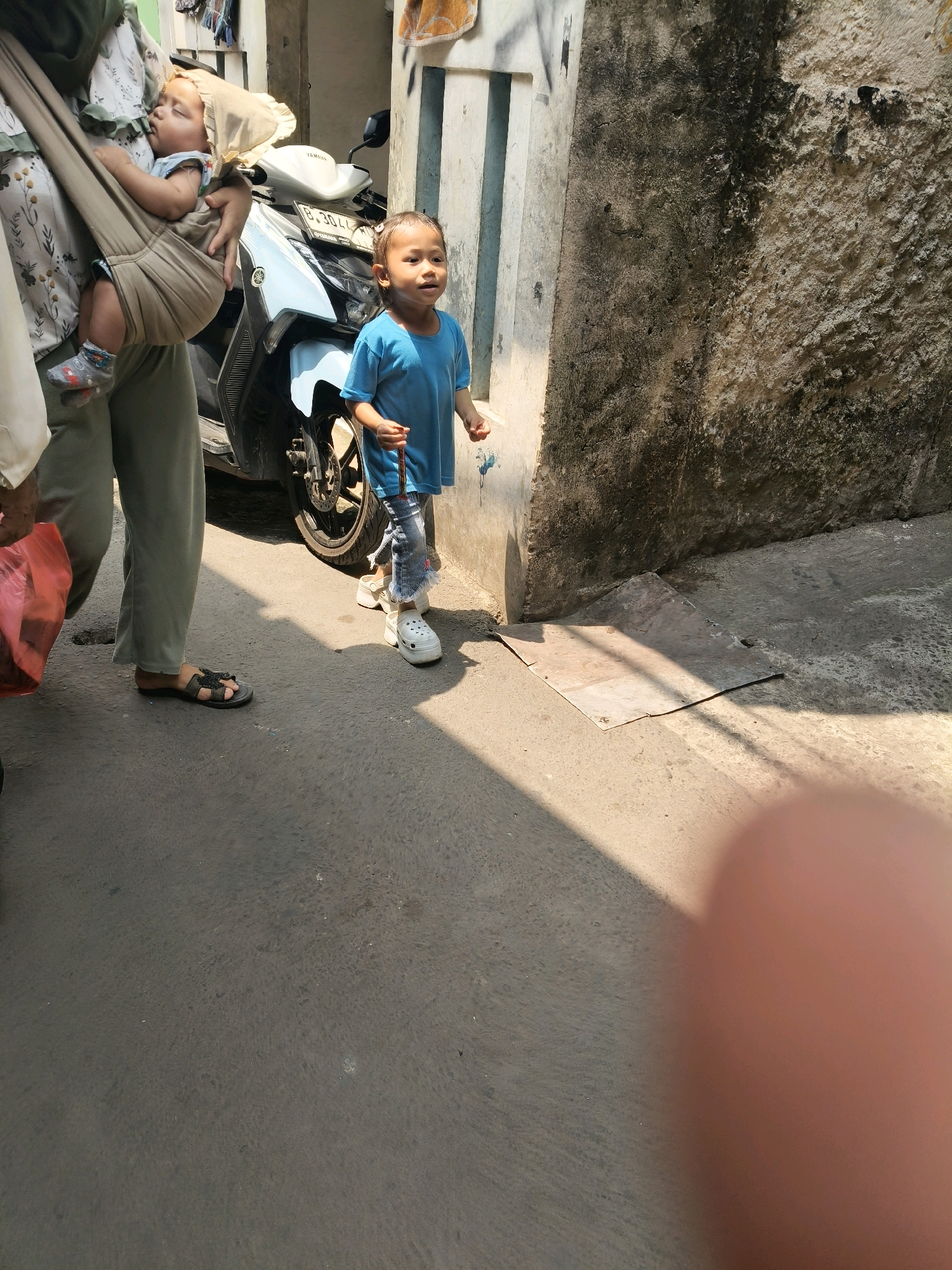 In South Tangerang, Indonesia, a toddler in a blue t-shirt and ripped jeans stands on a concrete street next to a motorcycle. The toddler is holding a thin object, possibly a stick or a toy. To the left, a person wearing a hijab and light-colored pants carries a baby in a sling. The baby appears to be sleeping, wearing a light-colored bonnet. The background features a textured, grimy wall and a portion of a building. The scene is illuminated by sunlight, casting shadows on the ground.