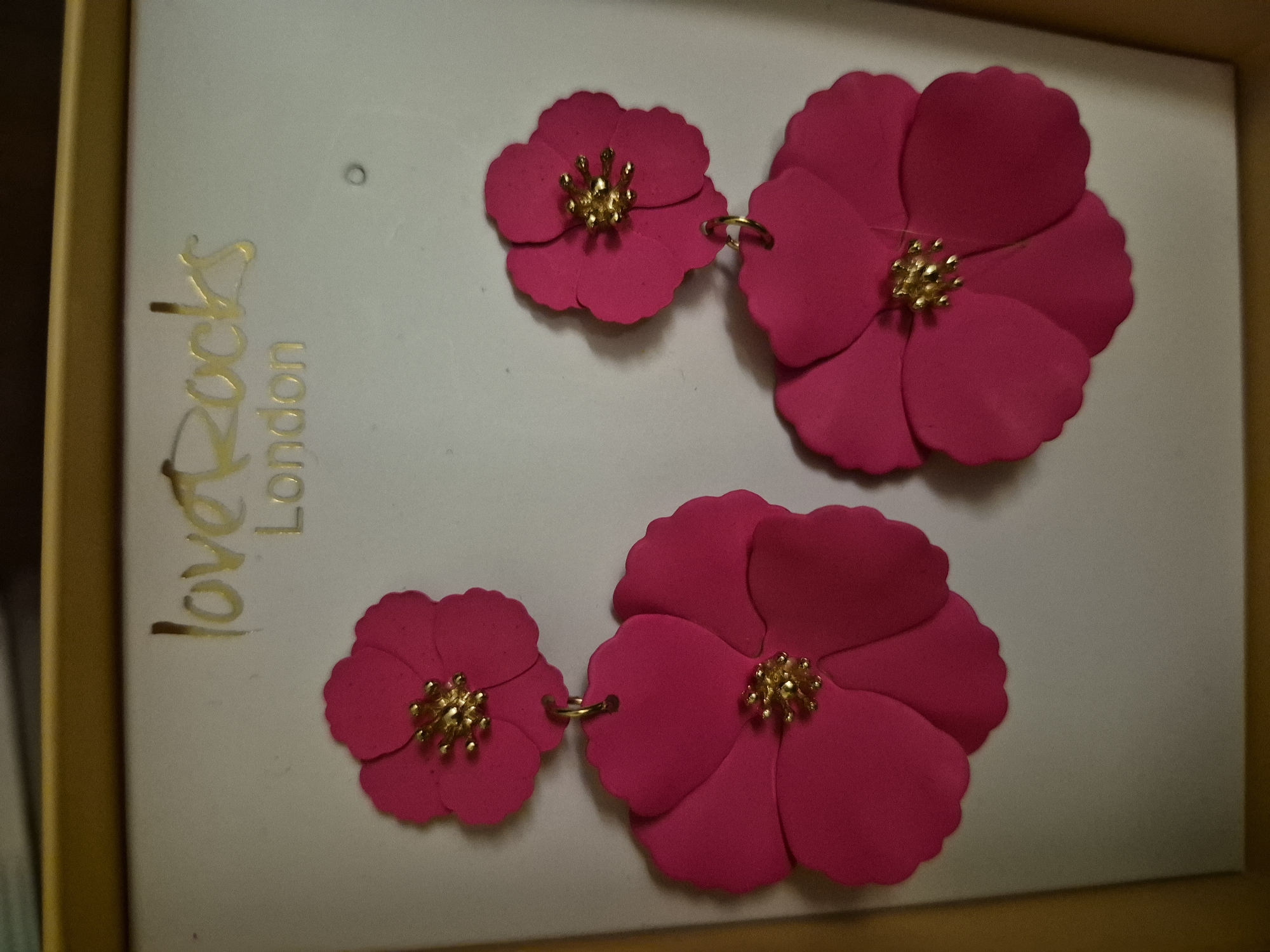 This is a close-up, eye-level, indoor, medium shot of a pair of bright pink, floral-shaped earrings displayed on a white background with gold lettering that reads "loveRocks London". The earrings are dangling, with a smaller flower on top and a larger flower below, both featuring gold centers. The background is a light gray color with a slight sheen, and a sliver of a gold border is visible at the top and bottom. The image appears to be taken in a studio or retail setting, with no signs of a specific city or urban environment.