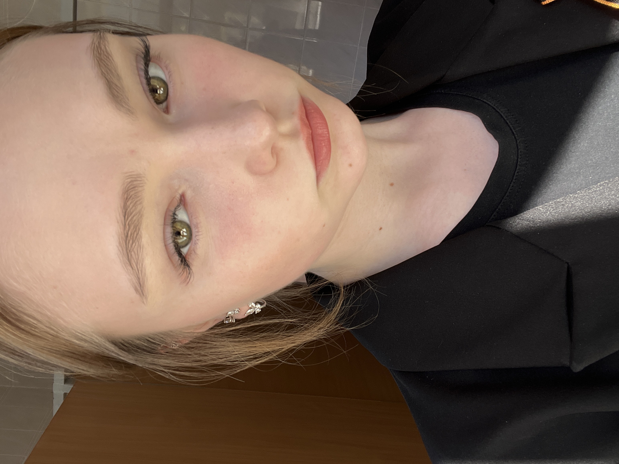A close-up, rotated portrait of a young woman with fair skin, light brown hair, and green eyes. She is wearing a black top and a pair of silver earrings. The background appears to be a tiled wall above a wooden surface. There is no indication or evidence that identifies a connection with the city of Chaikovsky, Russia.