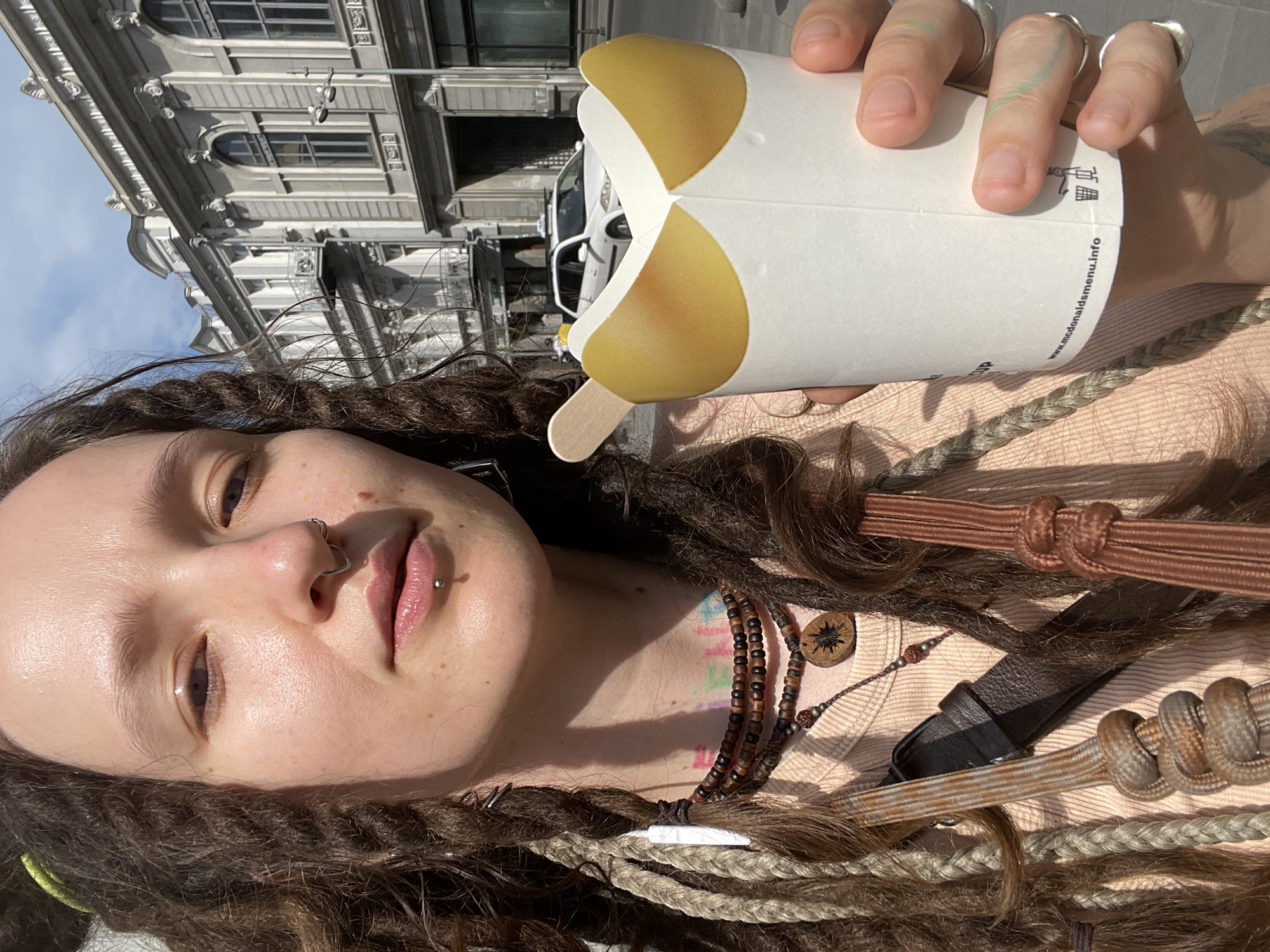 A person with dreadlocks, nose and lip piercings, and beaded necklaces is holding a white paper cup with yellow accents and a wooden stick. The person is wearing a light-colored shirt and appears to be outdoors in a city. In the background, there are buildings with architectural details and a white taxi. The scene is lit by sunlight. The bottom of the cup shows a website address and a recycling symbol. The location is identified as Tbilisi, Georgia.