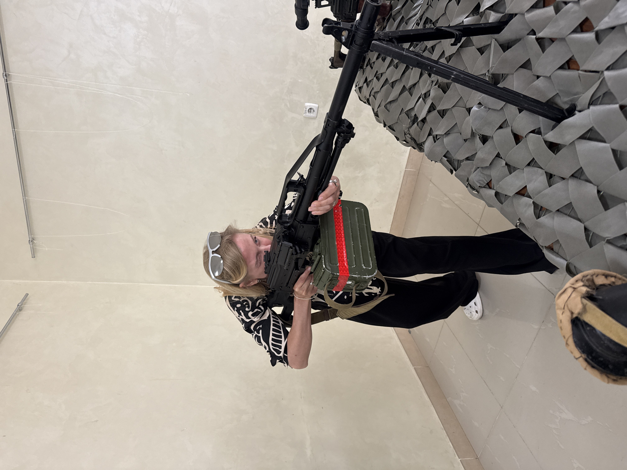 A woman stands holding a machine gun with a green ammunition box attached to it, aiming towards an unseen target. She is wearing sunglasses, a black and white patterned top, and black pants, with a white Croc sandal visible on one foot. The machine gun is mounted on a black metal stand which is covered with a gray camouflage net. The setting appears to be indoors, with a beige wall and a tiled floor.
