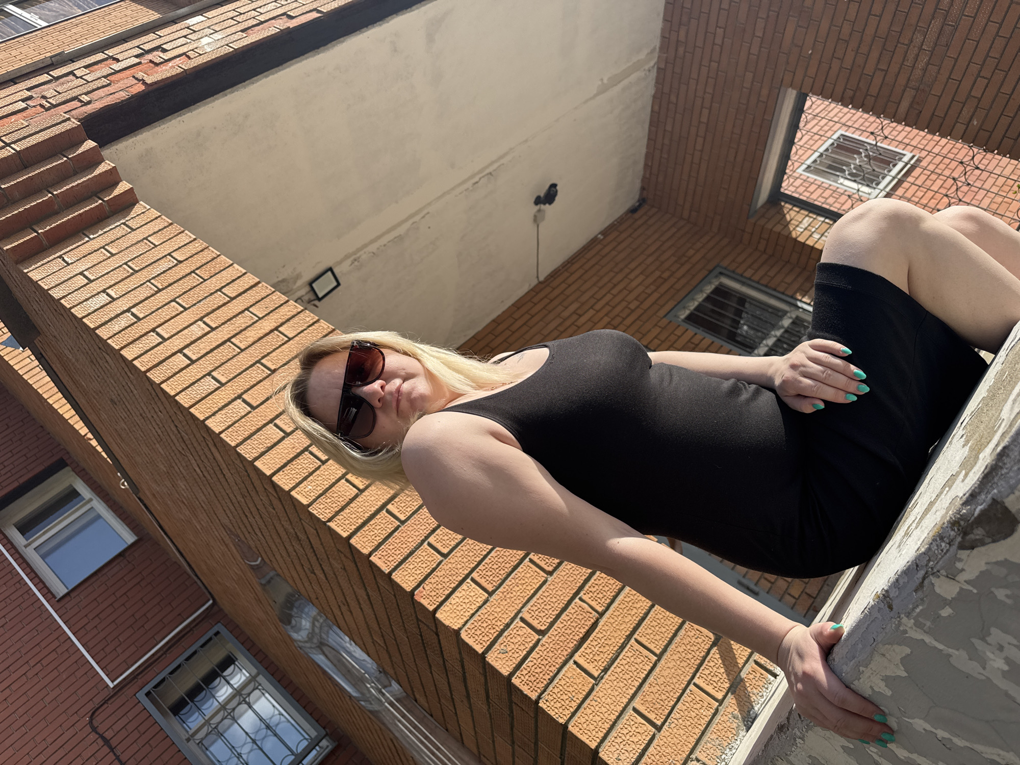 A low-angle shot reveals a woman posing on a low rooftop wall. She wears a black tank top and black shorts, and sunglasses, with blonde hair. Her nails are painted mint green. The background comprises brick apartment buildings, typical of urban Russia. The location is presumed to be in Chaikovsky, Russia, based on the request. The woman appears to be casually posing for the photo.