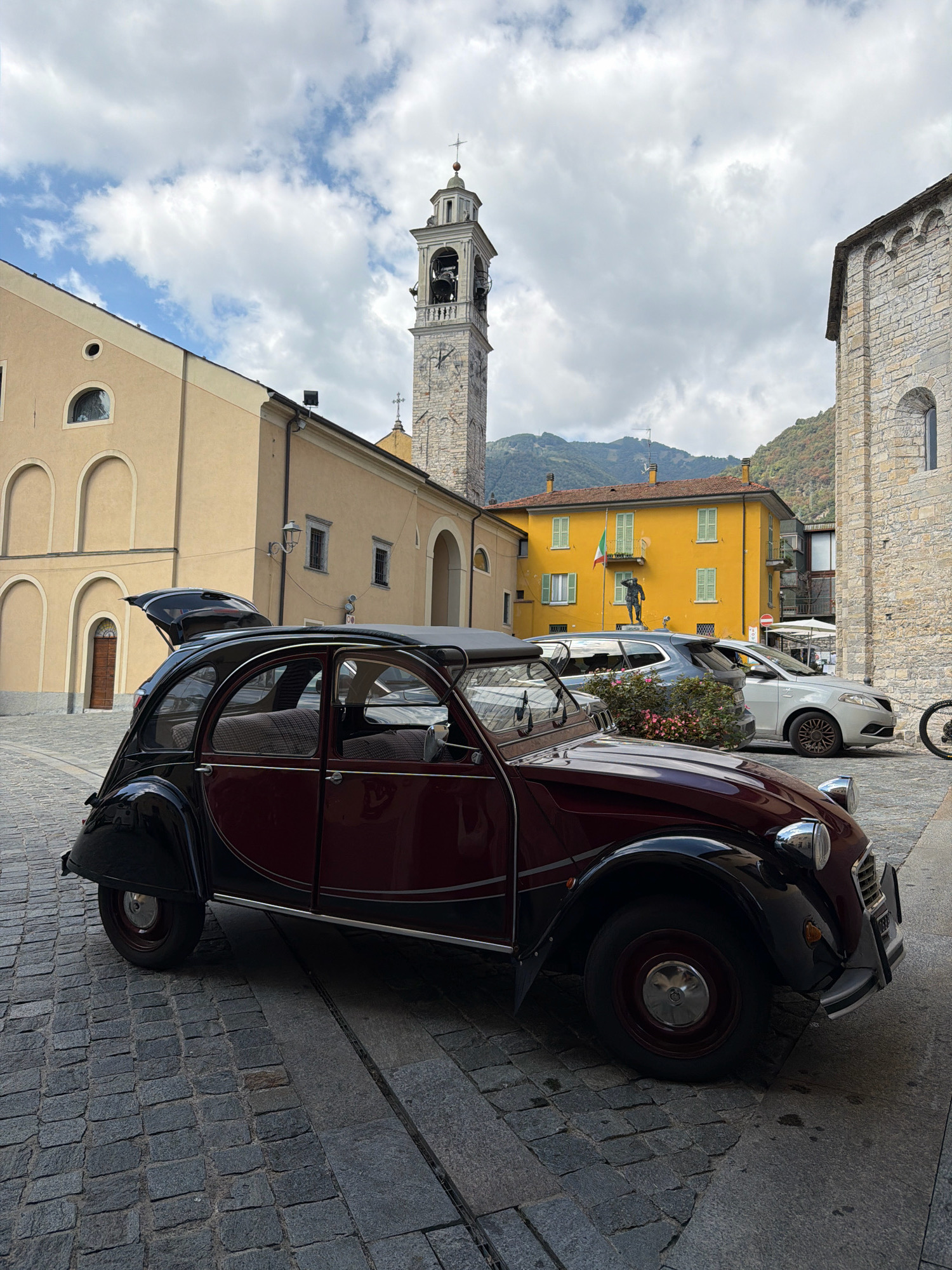 An old-fashioned, dark red and black two-door Citroën 2CV car is parked on a cobblestone street in front of a yellow building with arched windows and a beige building with arched entrances and windows. A tall stone bell tower with a clock face and arched openings stands behind the beige building, and a bright yellow building with green shutters and a statue in front is visible in the background, further back. A modern silver car and a dark grey SUV are parked behind the vintage car. The scene is set in Tremezzina, Italy, with a cloudy sky and a backdrop of green hills.