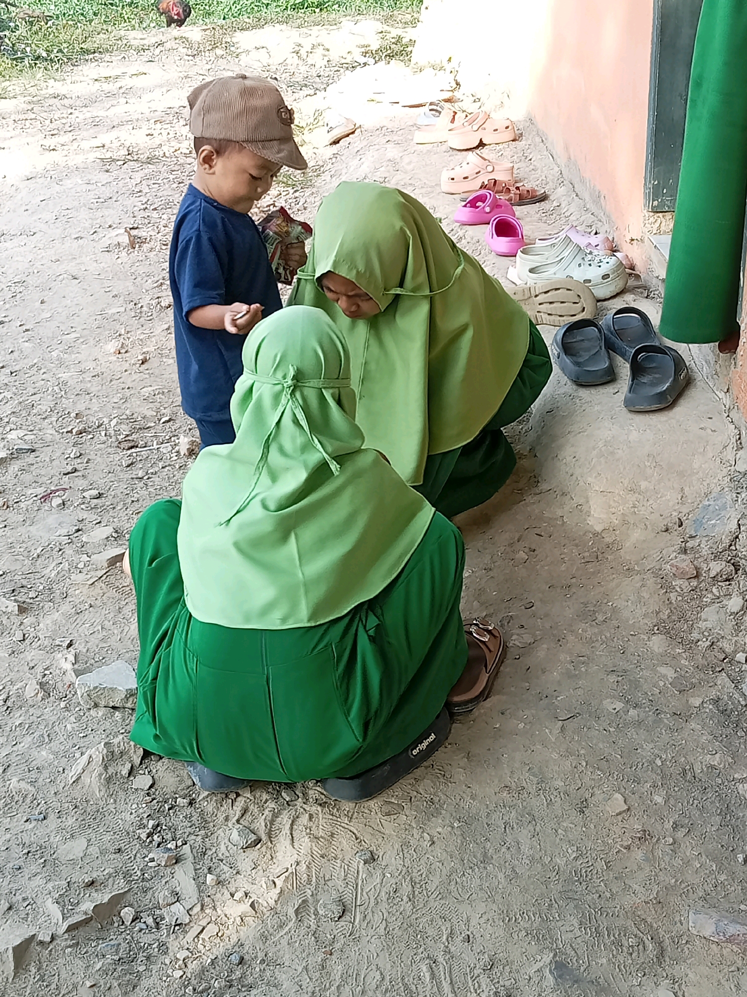 In Lebo Jaya, Indonesia, a young child wearing a blue t-shirt and a brown corduroy cap stands on a dirt path, holding a small object in his right hand. He is looking down. Two individuals wearing green hijabs are kneeling on the ground in front of him. The individual closer to the viewer has her back to the camera, and the other individual is facing the child. In the background, near a peach-colored wall, a collection of footwear is visible, including pink sandals, beige platform sandals, and dark gray clogs. A chicken is visible in the upper left corner of the image, amidst foliage. The ground is a mix of dirt, small stones, and debris, with visible footprints.