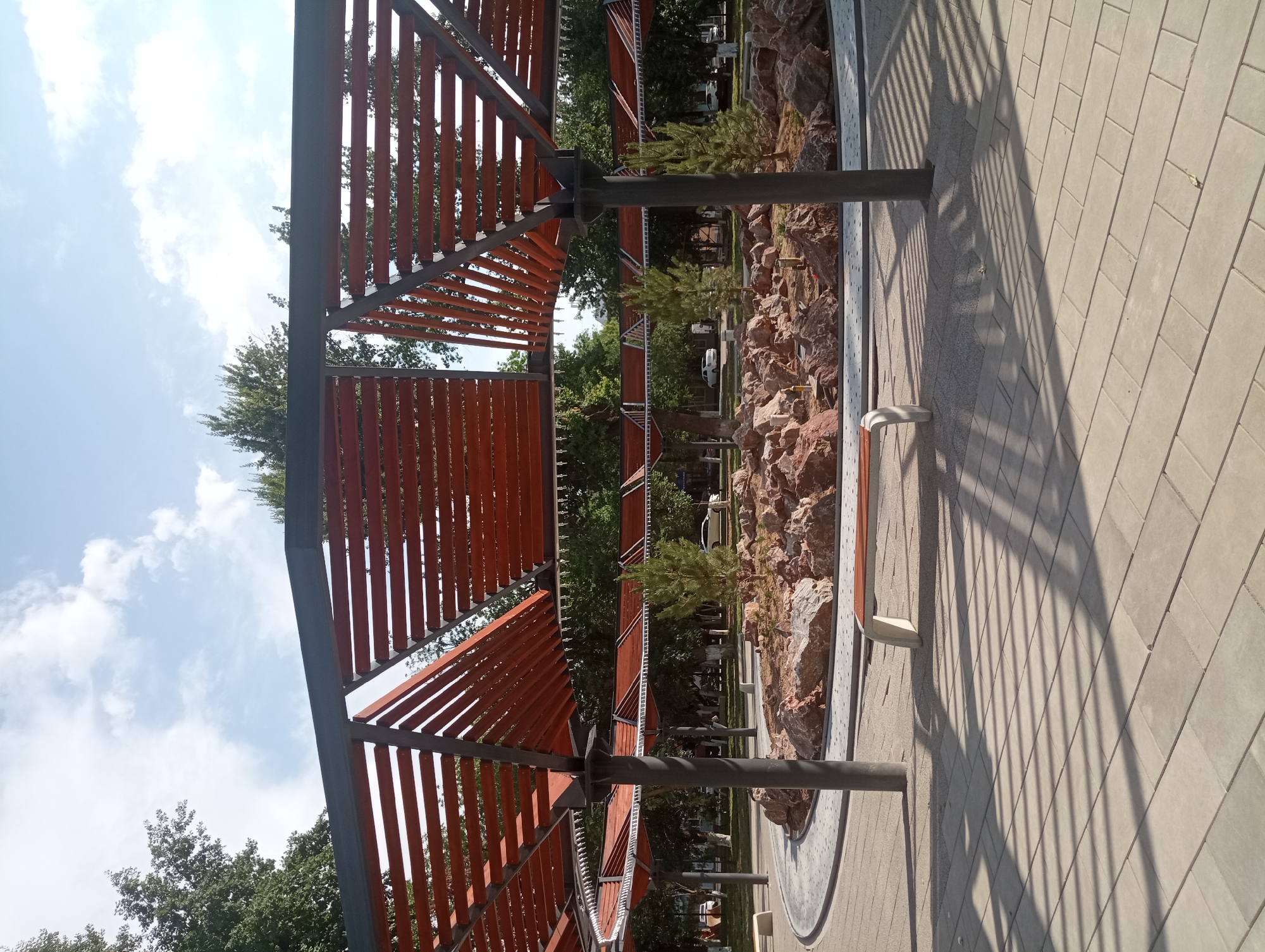 This media file depicts an outdoor scene in Shymkent, Kazakhstan, likely during daytime on a sunny day with some clouds. The main subjects are a modern pergola structure with a wooden slatted roof supported by dark metal beams, a decorative rock garden with small trees, and a paved walkway. The pergola casts a distinct shadow pattern on the walkway. A bench is partially visible within the rock garden area. In the background, there are more trees and glimpses of vehicles, suggesting a public park or a similar outdoor recreational space. The scene is tranquil and appears to be an architectural element designed for shade and aesthetic appeal within a naturalistic setting. No people are visible, and there is no visible text. The image is captured from a low angle, emphasizing the scale of the pergola and the play of light and shadow.