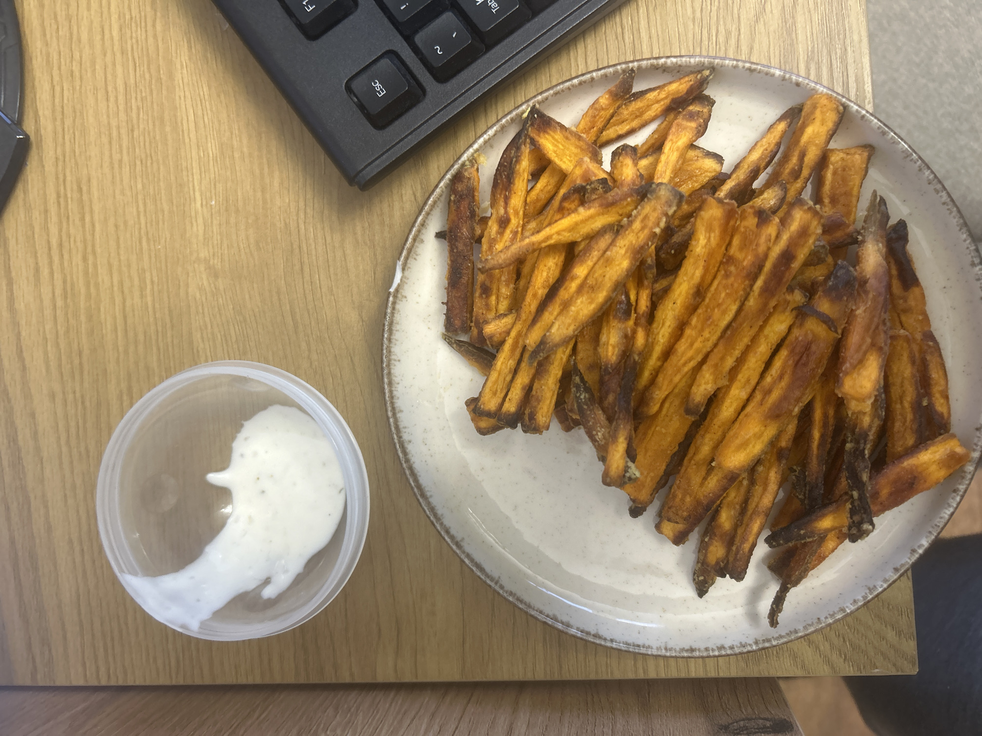 The image shows a plate of sweet potato fries next to a small container of dipping sauce and a black computer keyboard on a wooden table. The scene is indoors and it is not possible to determine if the image was taken in the city of Burgas, Bulgaria.