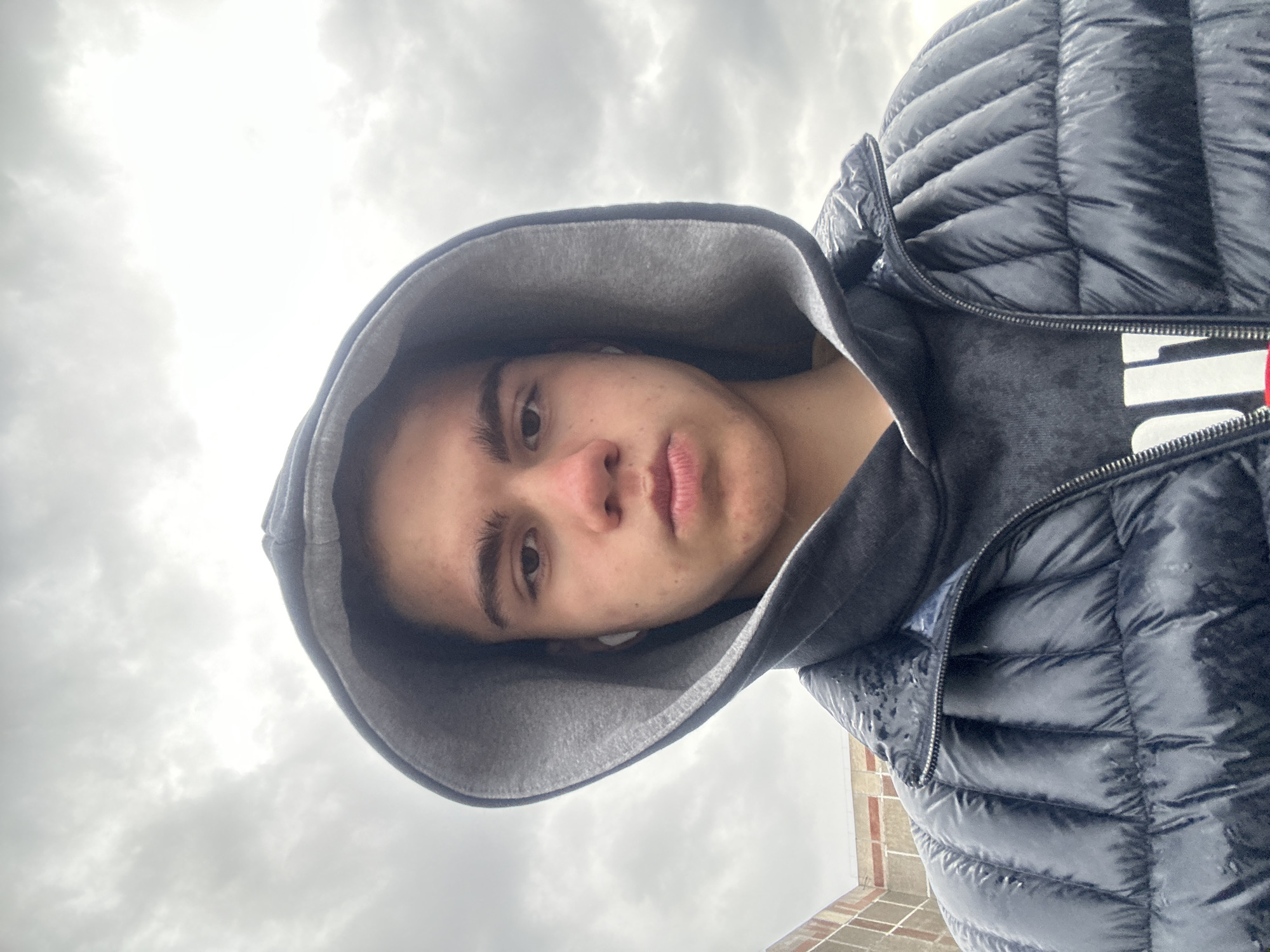 A young person with light skin and short brown hair is photographed from a low angle. They are wearing a black puffer jacket with a gray hood pulled up. The person is looking down at the camera. The background is a cloudy sky and a brick building. The location is the city of Unknown, Unknown. The photo appears to be taken outdoors during daylight.