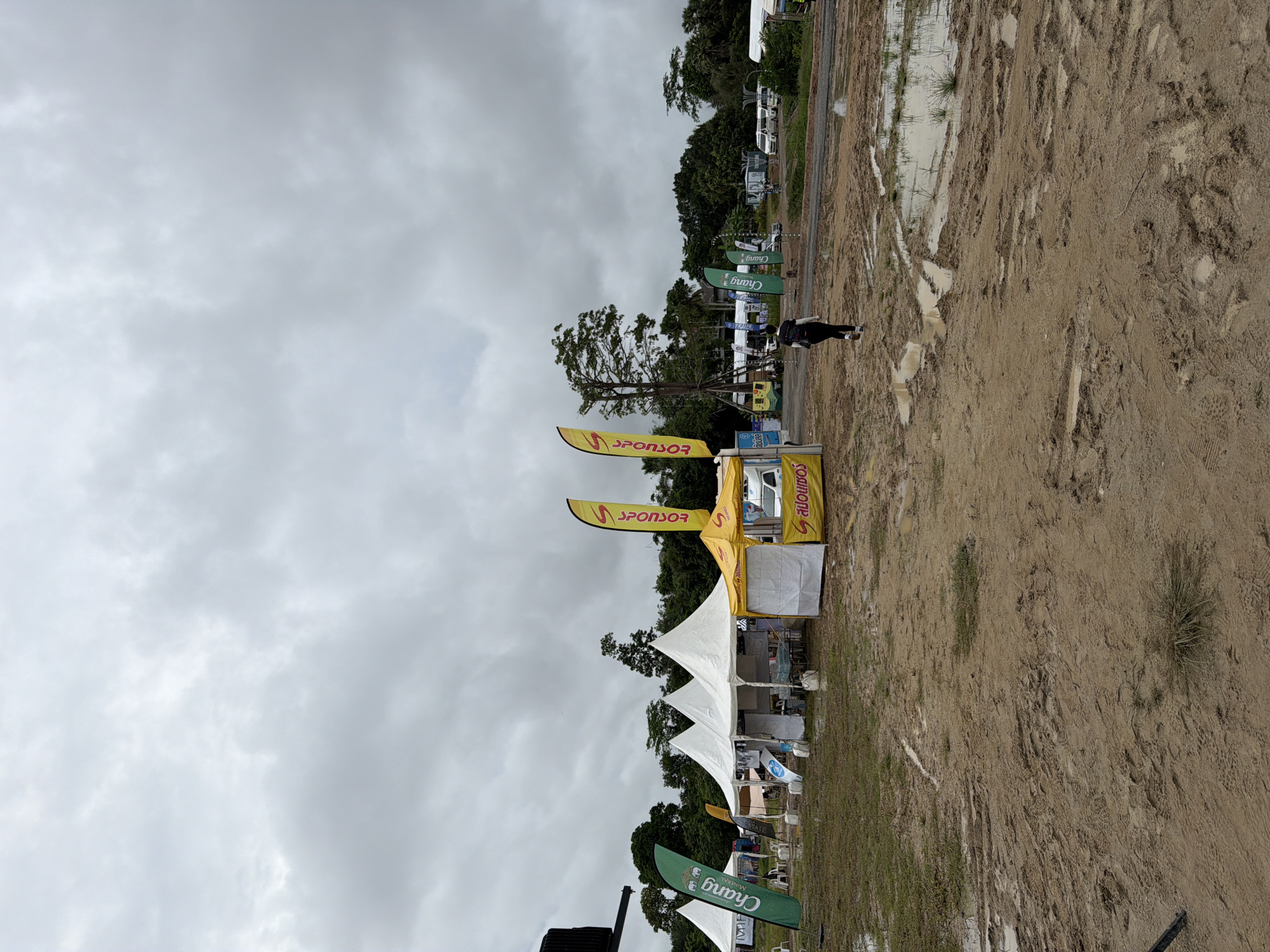 The image depicts an outdoor event, likely a festival or market, in Si Sunthon, Thailand, under a heavily overcast sky. The ground is muddy with standing water, indicating recent rainfall. Several white tents with peaked roofs are set up in the foreground, with a yellow tent on the right. Tall, slender flags with sponsor logos in yellow and green are planted near the tents. One flag on the right has the word "SPONSOR" in yellow text. A person is walking away from the camera towards the right, wearing black pants and a white top, with a backpack. In the background, there are trees and more tents, as well as several white vans parked on a paved area. The overall scene suggests a somewhat disorganized outdoor event, possibly affected by the weather.