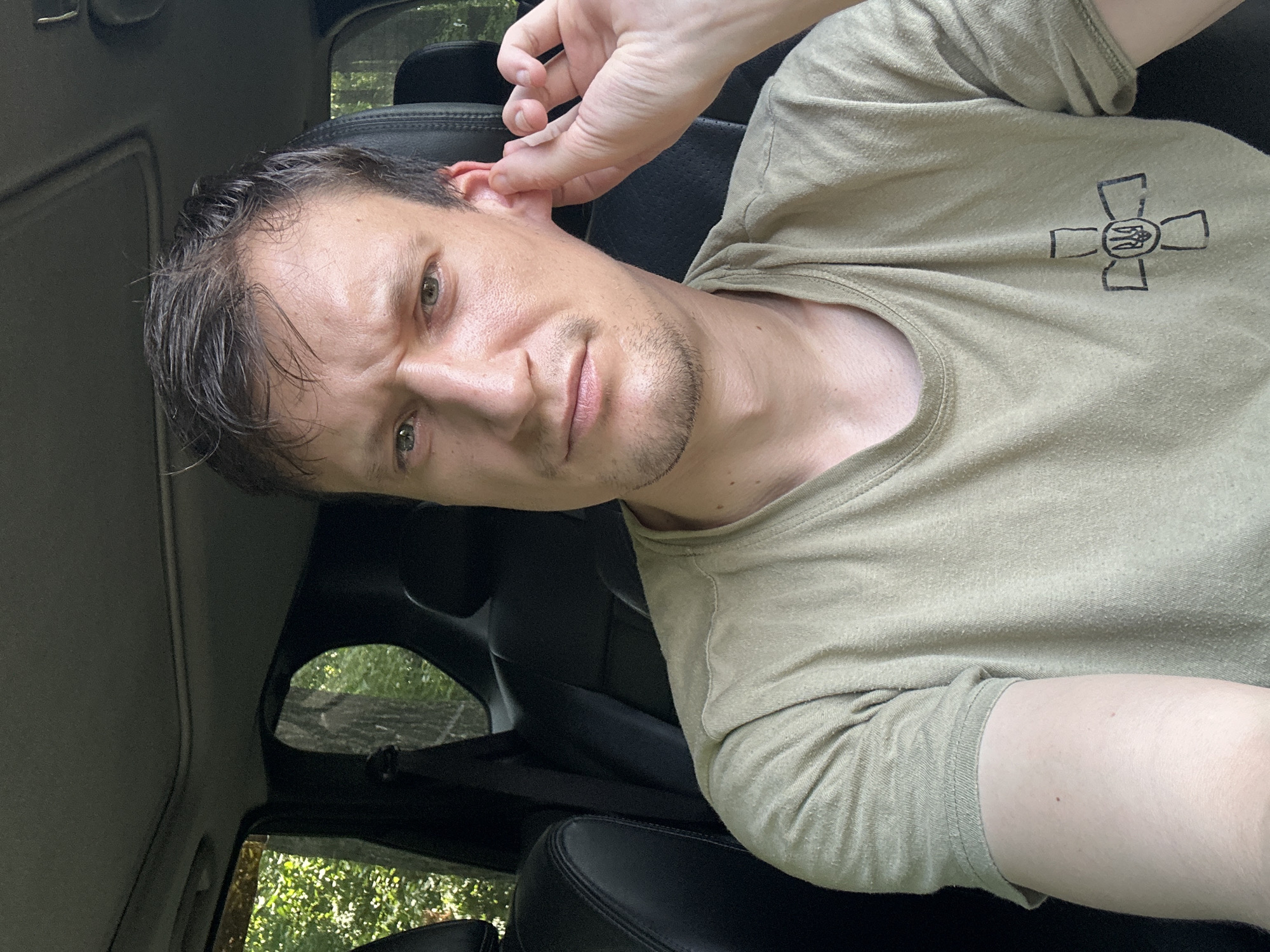 A man is sitting in the driver's seat of a vehicle, looking directly at the camera. His right hand is touching his right ear. He is wearing a light olive-green t-shirt with a black emblem on the left breast. The emblem is a cross with a shield in the center containing a trident. The interior of the vehicle is visible, including the black leather seats and part of the sunroof. Outside the vehicle, trees and foliage are visible, suggesting a location with natural surroundings. The man has short brown hair and a slight stubble. The lighting suggests it is daytime.