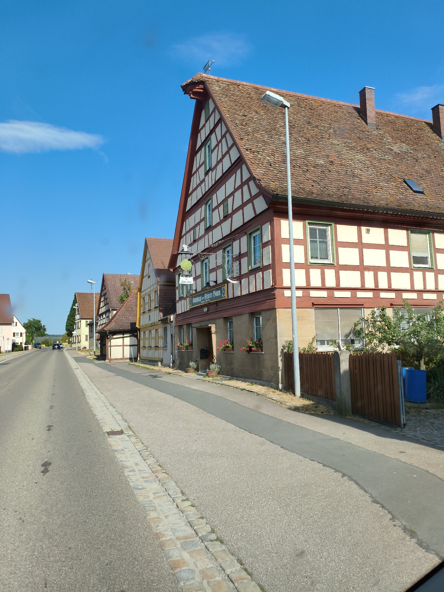 The image depicts a street scene in the city of Bad Windsheim, Germany, on a sunny day with a clear blue sky and a few wispy clouds. The street is paved, with a cobblestone border along the right side, separating it from a sidewalk. Several half-timbered buildings line the right side of the street, characterized by their red and white facades and red tiled roofs. One building prominently features a sign that reads "Gasthof Zum Grünen Baum" (Inn to the Green Tree). The buildings appear to be traditional German architecture. A lamppost stands on the sidewalk in front of one of the buildings. In the background, more buildings and trees are visible, suggesting a rural or suburban setting within the city. A car is seen driving away from the viewer down the street. The overall impression is one of a quiet, picturesque German town.