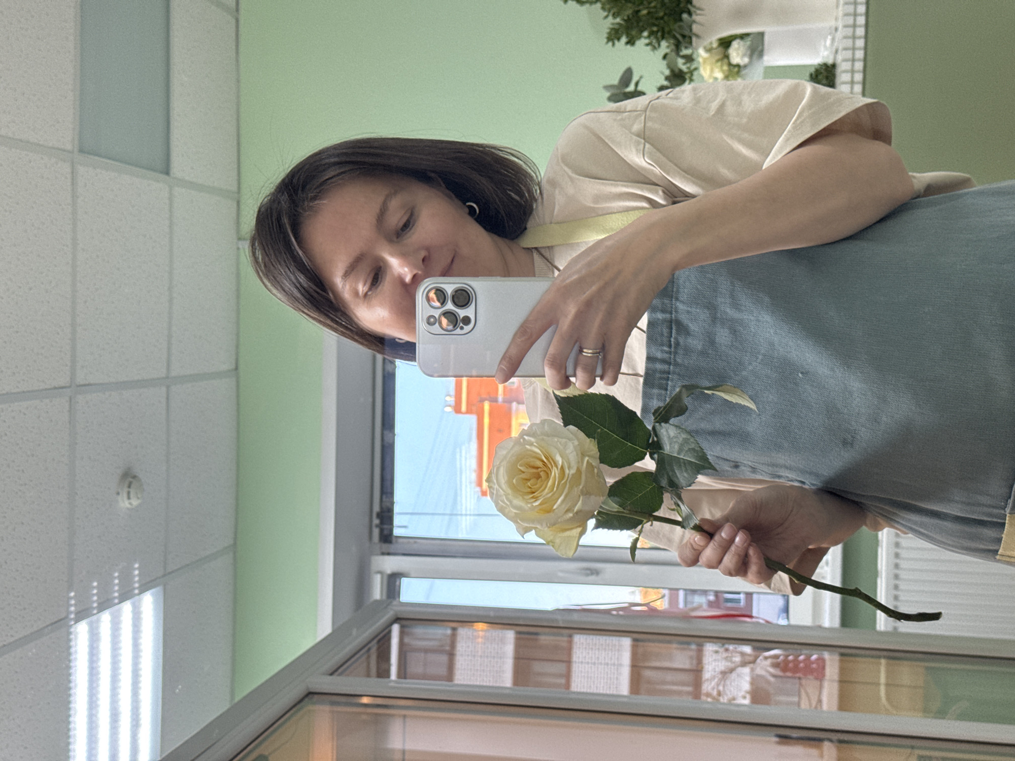 A woman is taking a selfie with a light yellow rose. The selfie is reflected in a glass window, and through the window, an orange building under a blue sky is visible. The building appears to be in the city of Izhevsk, Russia. The woman has dark hair, wears a light-colored t-shirt and blue denim. She is holding a white smartphone with multiple camera lenses. The background includes a ceiling with fluorescent lights, a green wall, and some greenery or potted plants. The image has been rotated.