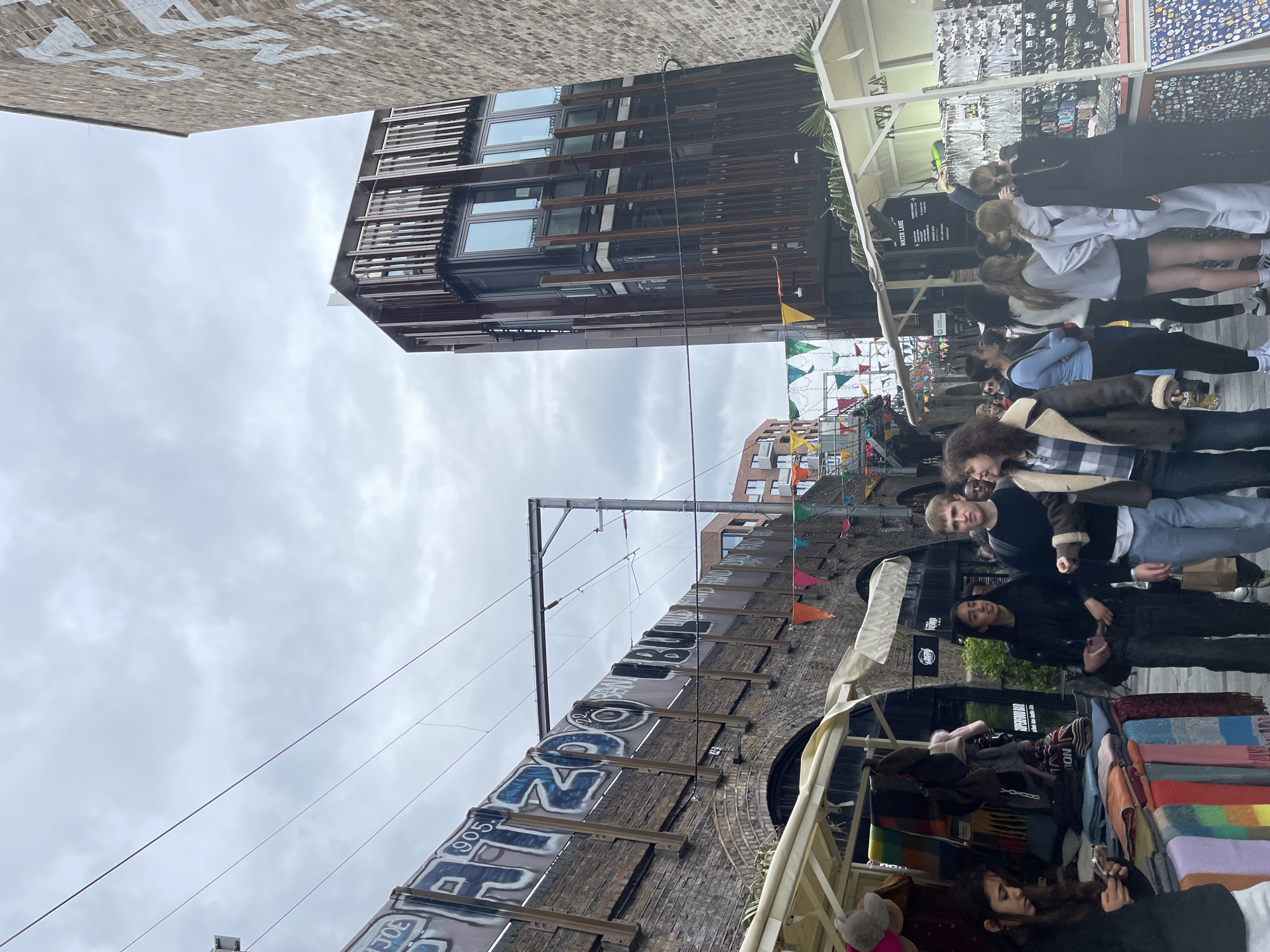 The image depicts a bustling outdoor market street, likely set beneath an elevated brick structure with archways in an urban environment under an overcast sky. Numerous people are seen walking, browsing, and standing around market stalls. In the foreground, people are casually dressed; a woman with dark curly hair is seen near a stall displaying colorful blankets and scarves, while a man with blonde hair and a beard looks towards the viewer. Further back, individuals are gathered around another stall that appears to sell jewelry and small items.
On the left, prominent brick archways are visible, featuring large, stylized graffiti with discernible letters such as "ON", "905", and "JOE". Overhead, electrical or tram lines stretch across the street. Colorful bunting, consisting of small flags, is strung between structures, adding a festive ambiance. On the right side of the street, a modern building with dark, slatted facades and large windows stands tall. A white sign with an arrow indicating "WATER LANE" is visible near one of the market stalls. Another partially visible sign reads "VINTAGE", and "RIVERFRONT" can be seen in the distance. The scene captures a lively daytime market activity.