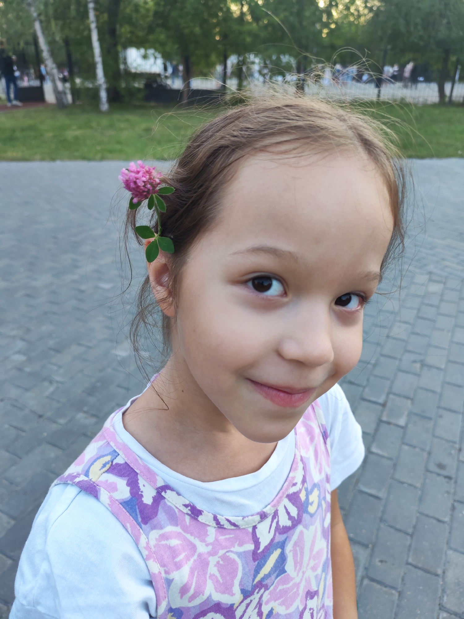 A medium shot captures a young girl with a pink clover flower tucked behind her right ear. She is looking to her right, with a slight smile on her face. Her dark brown eyes are open, and her hair is a light brown. She is wearing a white t-shirt layered under a purple and pink floral patterned dress. The background is blurred, showing a park setting with trees and a paved pathway made of grey rectangular stones. The lighting suggests it is daytime, possibly late afternoon. There is no visible text in the image.