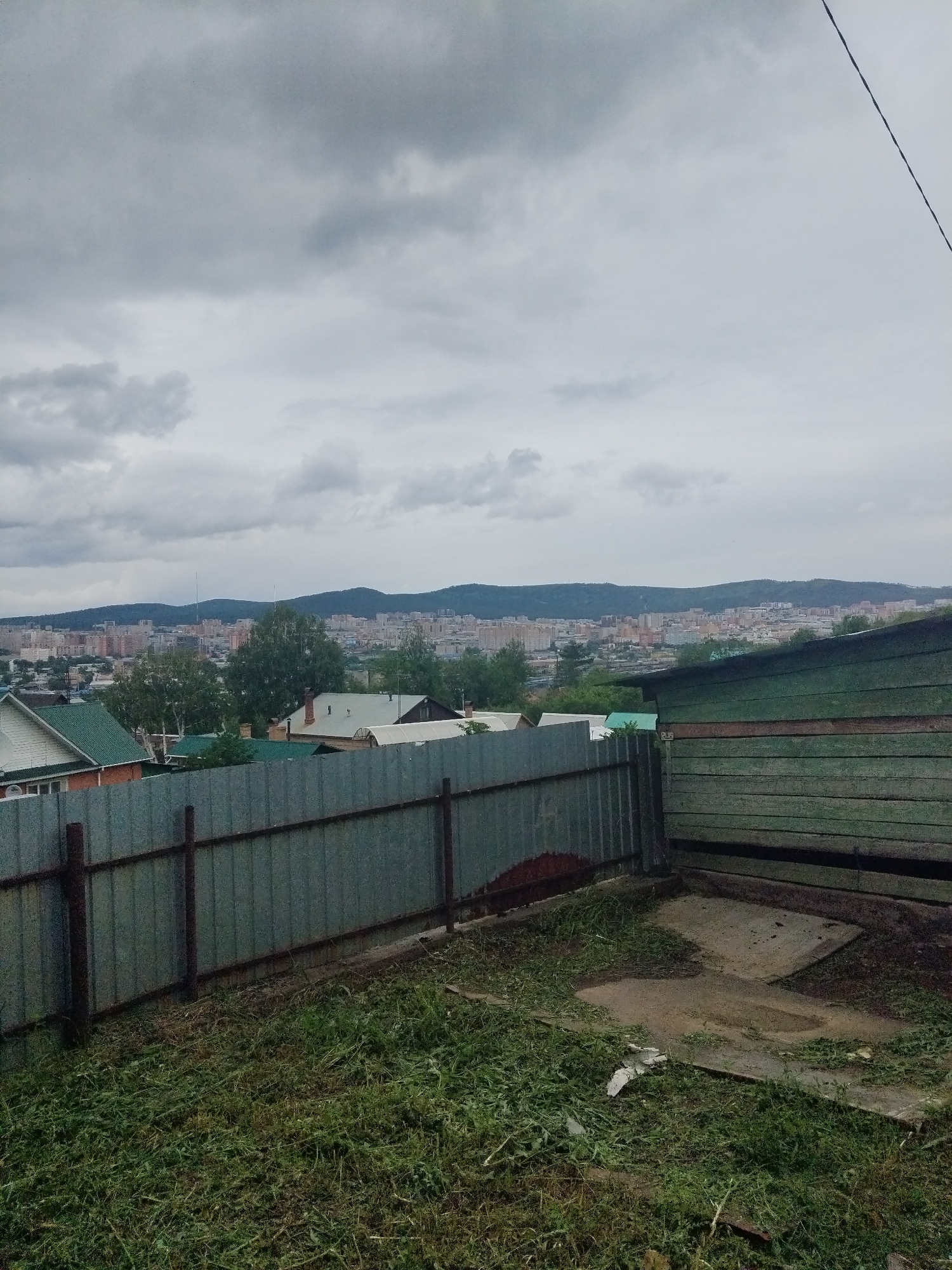 The image captures a view of Chita, Russia, from a slightly elevated position. In the foreground, a weathered green wooden structure, possibly a shed or outbuilding, is visible on the right, constructed with horizontal planks. Adjacent to it is a tall, corrugated metal fence, showing signs of rust and age, extending across the middle ground of the image. The ground in front of the fence is covered with freshly cut grass and some debris. Beyond the fence, a suburban landscape unfolds with houses featuring green roofs and other residential buildings. In the background, the city of Chita sprawls across the horizon, backed by rolling hills or mountains under a cloudy, gray sky. A single black wire stretches diagonally across the upper right portion of the frame.