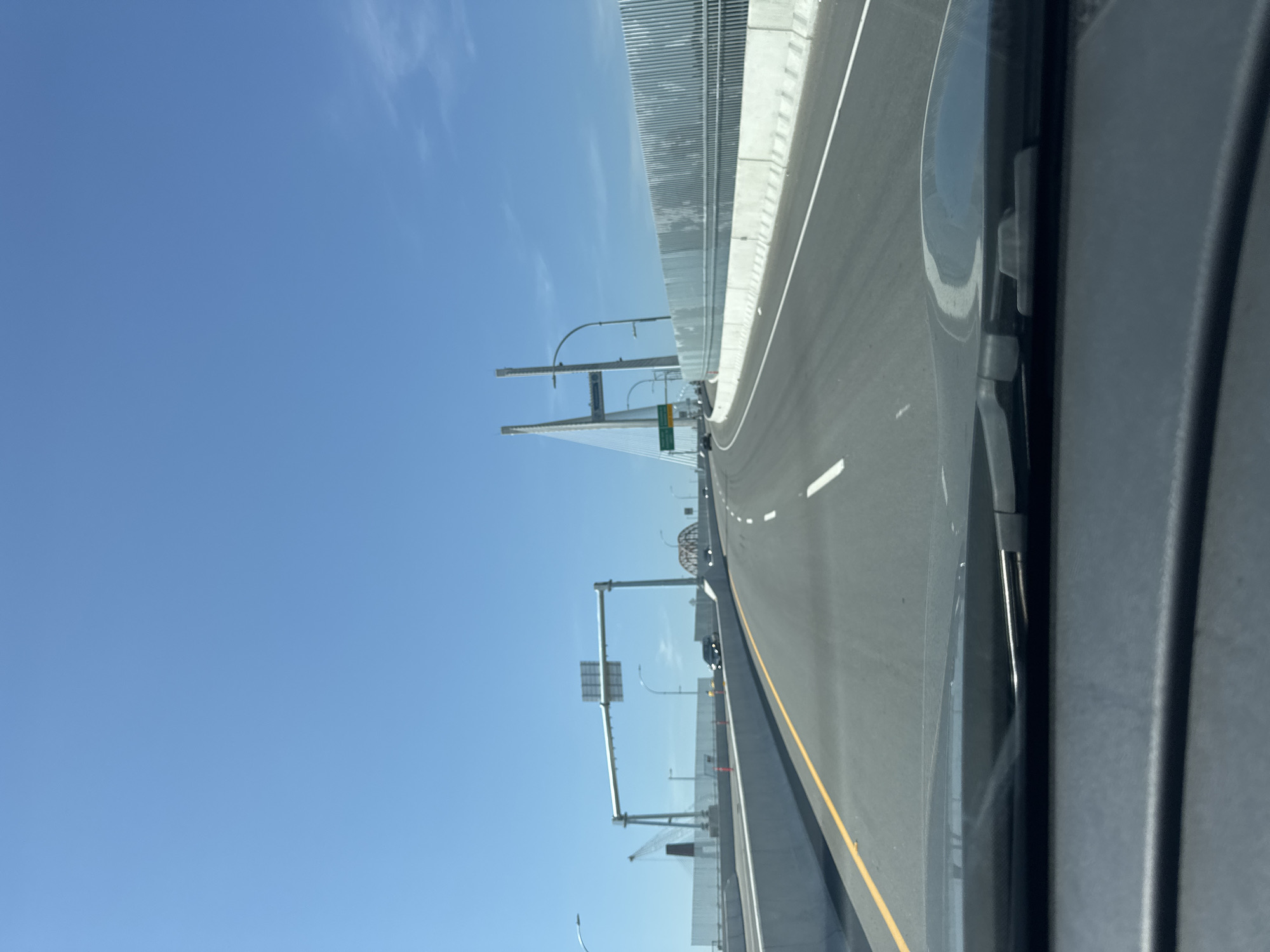 This is a point-of-view photograph taken from a vehicle traveling on a highway overpass or bridge. The sky is clear and blue, with a few wispy clouds. The highway is multi-lane and paved, with a yellow line marking the edge of the lane on the left. On the right side of the highway, there is a white concrete barrier topped with a grey, corrugated metal fence. In the distance, a large suspension bridge with two prominent white towers can be seen. There are also traffic lights, signs, and other vehicles visible on the road. The foreground shows the dashboard and windshield wipers of the vehicle. Based on the presence of the cable-stayed bridge and highway infrastructure, this image likely depicts a section of the Port Mann Bridge or the Highway 1 expansion project in Surrey, British Columbia, Canada.