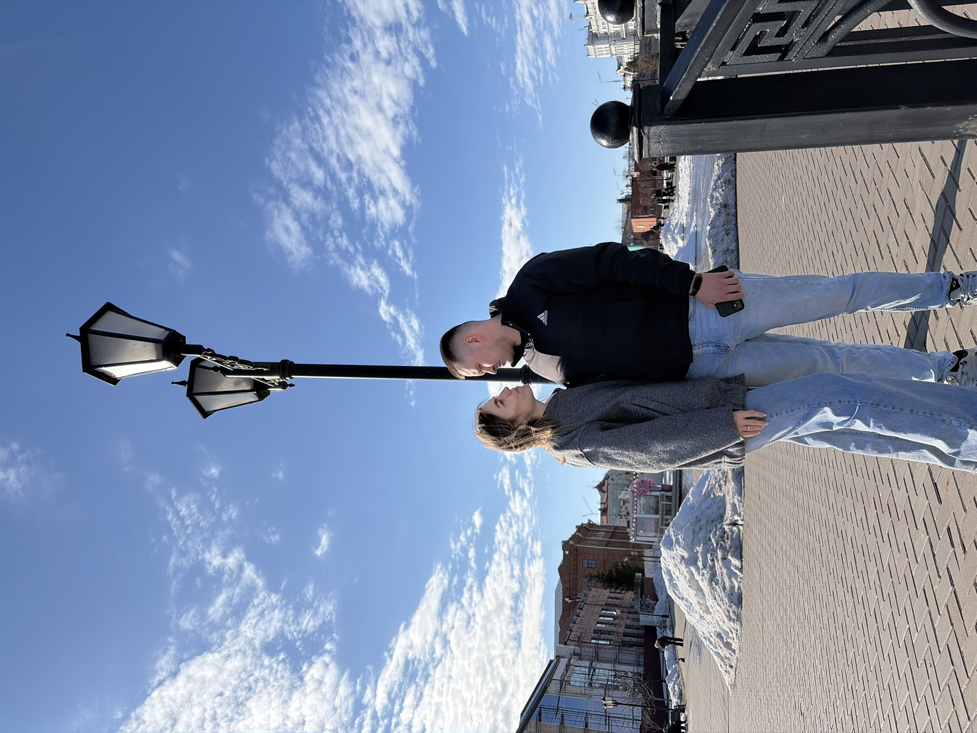 A couple is standing face-to-face on a brick walkway in Sarapul, Russia. The man is wearing a black jacket and light blue jeans, and holding a cell phone. The woman is wearing a grey coat and light blue jeans. A black lamp post is to their left, and a black iron railing is behind the man. Buildings are visible in the background, as well as patches of snow. The sky is blue with wispy clouds.
