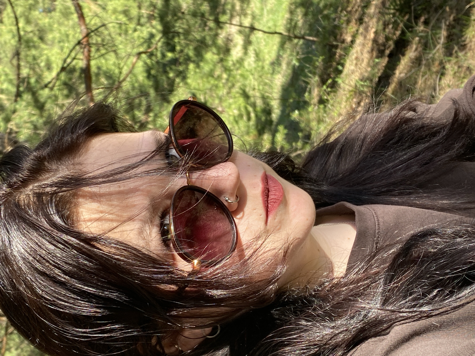 A young woman with dark hair and sunglasses is lying in a grassy, wooded area. Her face is turned towards the viewer, though her head is tilted to the side. She is wearing a brown top. The background shows green grass and trees, and the overall impression is of a sunny day outdoors. The location cannot be determined from the image; it is not possible to verify the city of Фоки, Russia.