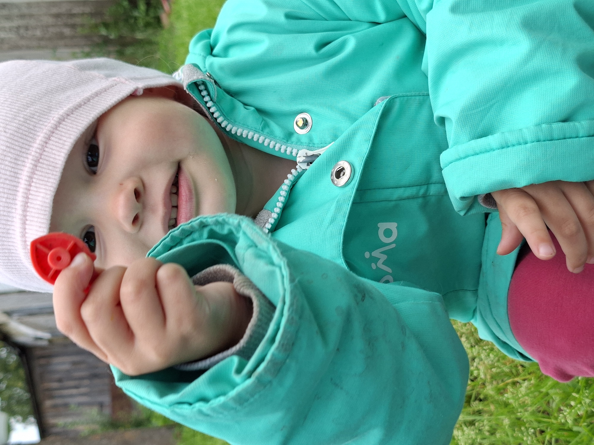 The image shows a close-up, slightly tilted view of a young child wearing a pink hat and a turquoise jacket. The child is holding a small red object in their right hand, which appears to be a toy or a part of a toy. The child's face is partially visible, with a smile and bright eyes. The background is blurred, showing green grass and some wooden structures, suggesting an outdoor setting, possibly a backyard or a park. The lighting suggests it is daytime. The turquoise jacket has a zipper and buttons, and a white trim around the collar. There are some small dark marks on the jacket, possibly dirt or stains. On the jacket, there is visible text that appears to be a logo or brand name, reading "REIMA" in white letters. The child's expression is joyful and engaged. The image is a candid shot, capturing a moment of play.