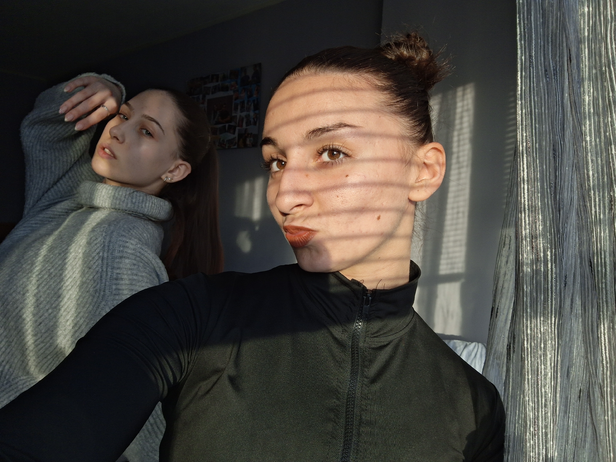 The image depicts two young adult females posing indoors in Sofia, Bulgaria, illuminated by sunlight casting distinct shadows.
The foreground subject, positioned right of center, has dark hair tied in a bun and brown eyes. She wears a black, high-collared, zippered long-sleeved top and reddish-brown lipstick. Her face is turned towards the camera with a slight pout, and horizontal light stripes, consistent with shadows from window blinds, fall across her face.
The background subject, positioned left of center, has light brown hair in a ponytail and light-colored eyes. She wears a light grey knitted turtleneck sweater. Her head is tilted, and she looks towards the camera over her left shoulder, with her left hand raised near her head. A silver ring is visible on her left ring finger.
The indoor environment features a dark grey wall in the background to the left, adorned with a grid of multiple small rectangular images. To the right, behind the foreground subject, a silvery-grey curtain with vertical stripes is visible, suggesting a window covering. The strong directional sunlight creates a series of parallel shadows on the subjects and the walls, indicating light entering from a window with a slatted covering.