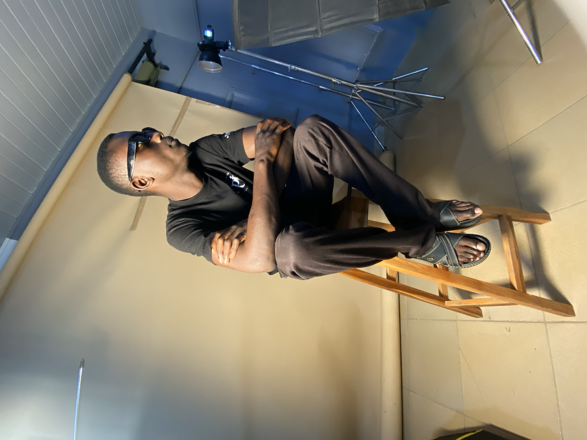 A young man sits on a wooden stool with his arms crossed and his legs crossed, wearing a black t-shirt, dark pants, and black sandals. He has sunglasses on and is looking off to the side. The background appears to be a studio setting with a cream-colored backdrop and some photography equipment, including a light stand with a light and a softbox. The floor is tiled. The overall lighting suggests an indoor studio environment.