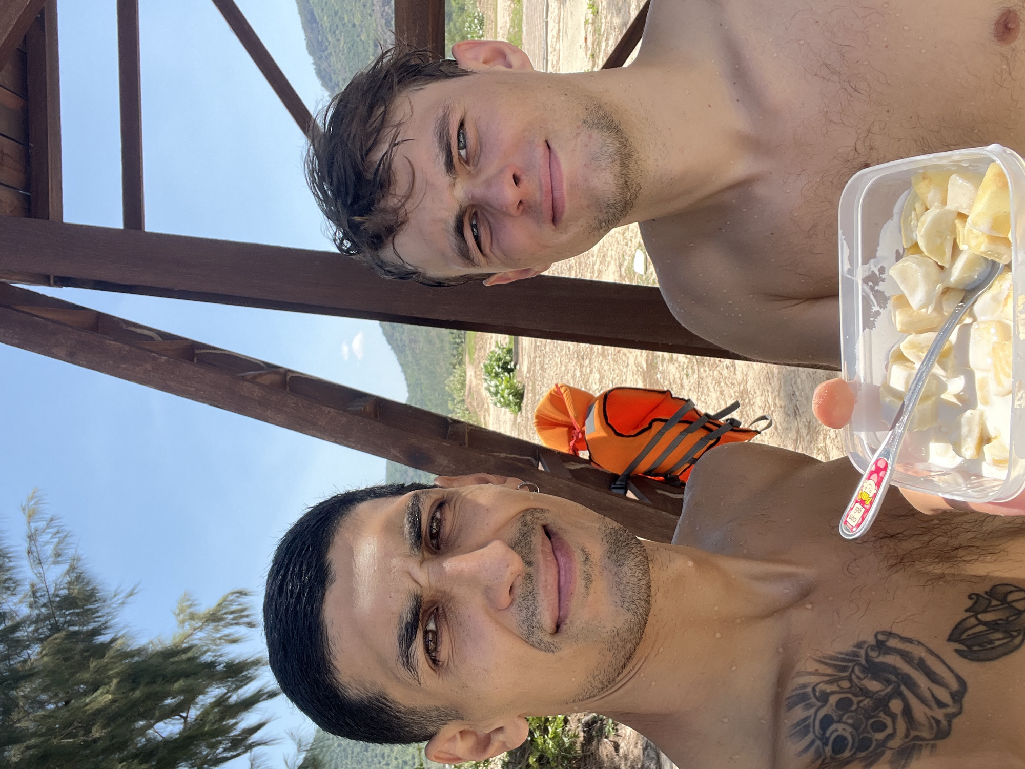 Two men are captured in a close-up outdoor selfie, presumably in Cong Hai, Vietnam, under a clear blue sky. The man on the left, with dark, wet, short hair and a light beard, wears a small hoop earring on his right ear and displays multiple tattoos on his bare chest, including a prominent design of what appears to be a skull or similar figure holding guns on his left pectoral. He holds a clear plastic container filled with diced fruit (predominantly bananas) mixed with a white creamy substance, a small spoon depicting a cartoon character, and a small orange spherical fruit resting on the container's edge. His expression is a slight smile. The man on the right, with brown, wet, messy hair and light stubble, is also bare-chested and looks at the camera with a slight smile. Water droplets are visible on both men's skin. In the mid-ground, an orange and grey life vest hangs on a dark brown wooden structure that dominates the right side of the frame. The background reveals sandy or dirt ground with sparse greenery, leading to large, tree-covered mountains under a bright, sunny sky.