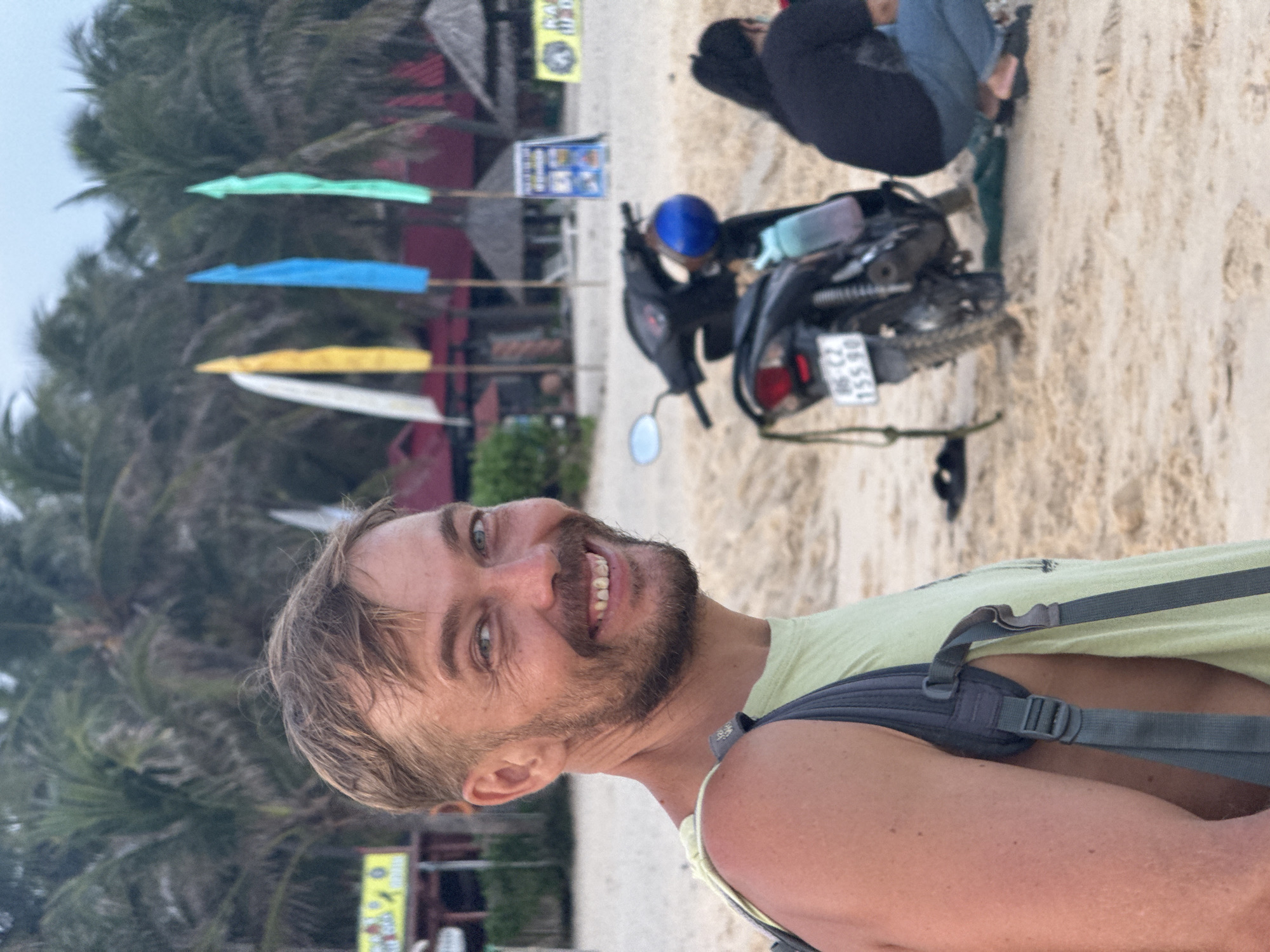 A man with a short beard and light brown hair, wearing a light green tank top and a grey backpack, is smiling and looking at the camera on a sandy beach. In the background, there are palm trees, several colorful flags, a black scooter, and two people in the distance, one crouched down. The scene suggests a tropical or coastal location, possibly Phu Thuy Ward, Vietnam, given the presence of palm trees and beachside establishments.
