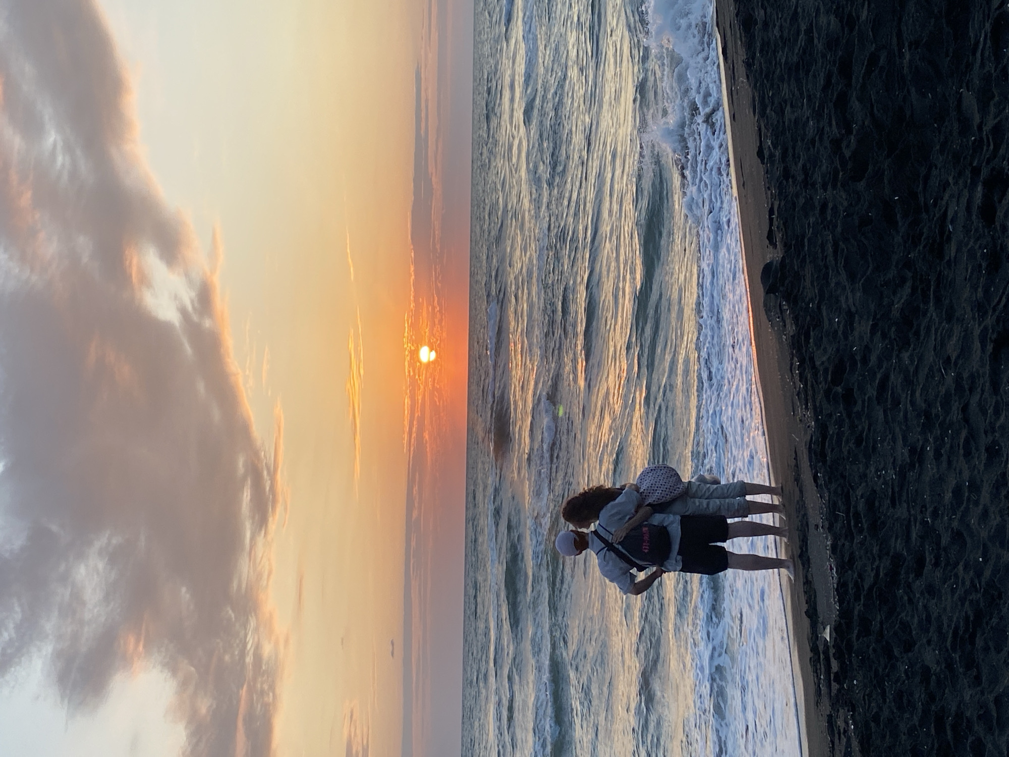 A couple stands facing the sea on a beach in Ureki, Georgia, during sunset. The man, wearing a light-colored shirt and dark shorts, has a backpack slung over his shoulder. The woman, beside him, wears light-colored pants and a light-colored top, also with a bag. They are positioned on dark sand, with the waves of the sea gently lapping at the shore. The sky is filled with pastel-colored clouds, illuminated by the setting sun, which casts a warm glow over the scene. The sea exhibits a moderate swell, with whitecaps visible as the waves break. The horizon line is clearly defined, separating the ocean from the sky. The overall atmosphere is serene and romantic, characteristic of a sunset by the sea.