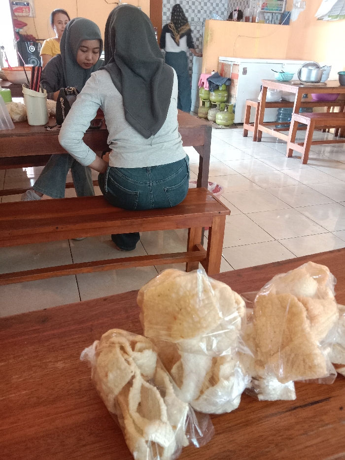 A restaurant interior with a table laden with bagged crackers in the foreground. Two women wearing hijabs sit at a table, one in a grey top and denim pants. Another woman stands near the back. In the background are a white refrigerator, gas tanks, another table, and tiled walls. The floor is tiled.