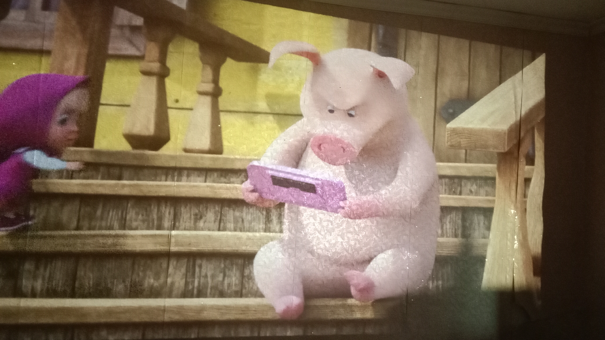 The image shows a scene from an animated production, likely projected onto a screen, depicting two cartoon characters. On the left is a small character, appearing to be a child, wearing a purple hood and a light blue shirt, reaching out to grasp onto wooden steps. To the right, a large, pink pig character with an angry expression is seated on the same wooden steps. The pig is holding a rectangular pink device with a screen, possibly a video game console, in its trotters. The background consists of wooden structures, including railings and vertical posts, with a yellowish-green wall behind them. The overall environment suggests an outdoor setting, perhaps a porch or a farmyard, rendered in a cartoonish style. There is no discernible location in Petropavl, Kazakhstan depicted in this image.