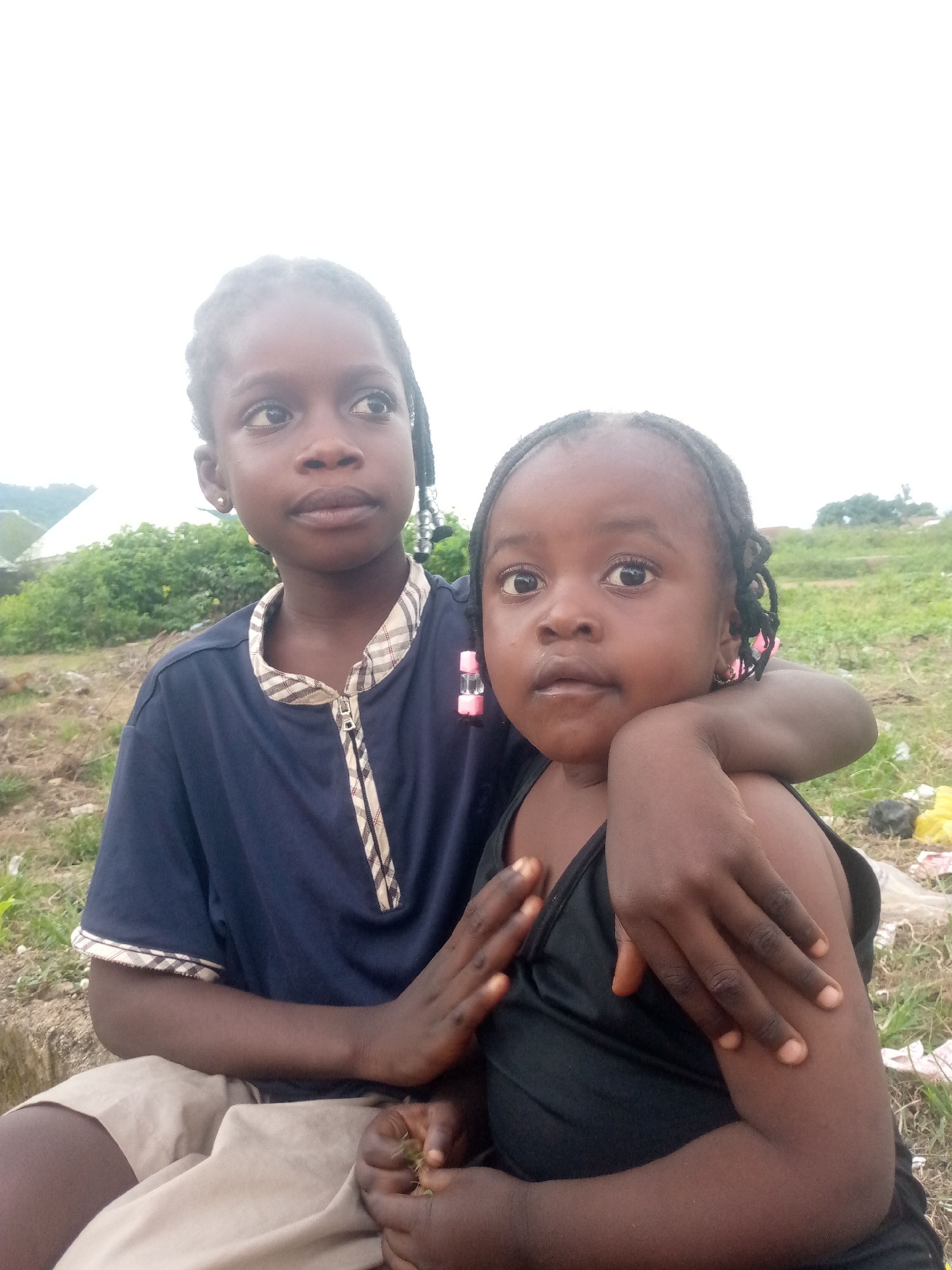 Two young Black girls are posed in a close-up portrait outdoors, with the older girl on the left and the younger girl on the right. The older girl, with braids adorned with black and silver beads, wears a dark blue collared shirt with a plaid trim. Her head is turned slightly to the left, her eyes looking upwards. The younger girl, her hair in braids with pink beads, is looking directly at the camera, wearing a black tank top. Her older sister's arm is around her shoulders, and the younger girl's arm is resting on her sister's arm. They are seated on the ground in a grassy area, with a slightly overcast sky in the background. Some vegetation and what appears to be refuse are visible in the background, suggesting a semi-rural or undeveloped urban setting in Serti, Nigeria.
