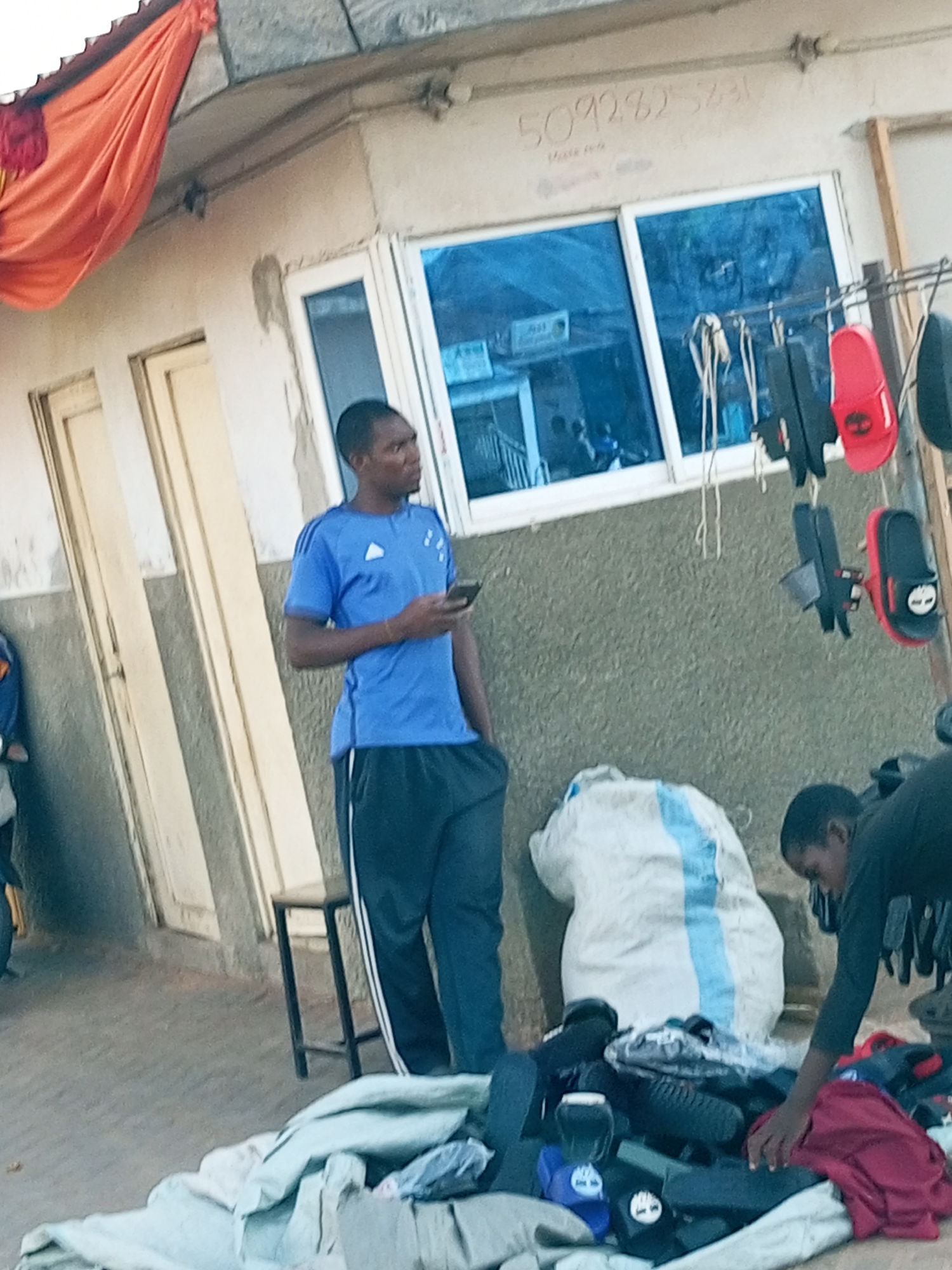 A man in a blue t-shirt and black pants is standing and looking to the right while holding a dark smartphone. He is positioned in front of a building with a blue window. Next to him, to his right, a rack holds various sandals and flip-flops. In the foreground, a pile of clothes and bags is visible. Another person, wearing a dark shirt, is bent over a pile of items to the right of the frame. The scene appears to be an outdoor market or storefront in Gombe, Nigeria, indicated by the presence of street vendors and the architectural style. The building has a textured, grey wall and a white framed window. An orange fabric is draped from the top left of the building. There is some writing on the wall above the window, but it is illegible.
