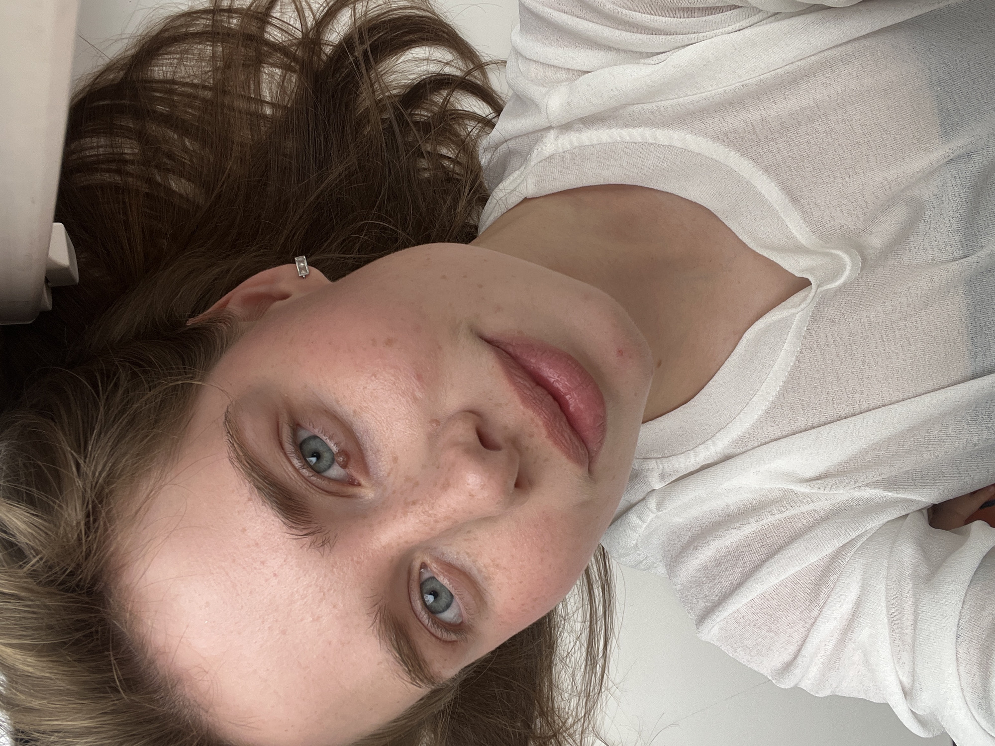 An overhead, medium shot captures a young Caucasian woman lying on a flat, white surface, possibly a desk or table. She is wearing a sheer white top. Her head is tilted to her right, and her face is turned towards the camera, with her brown hair spread around her head. She has fair skin with some freckles across her nose and cheeks, light pink lips, and light blue eyes. A silver hoop earring is visible in her left ear. The image appears to be taken indoors in a residential or studio setting, with no clear indication of the city of Chaikovsky, Russia.