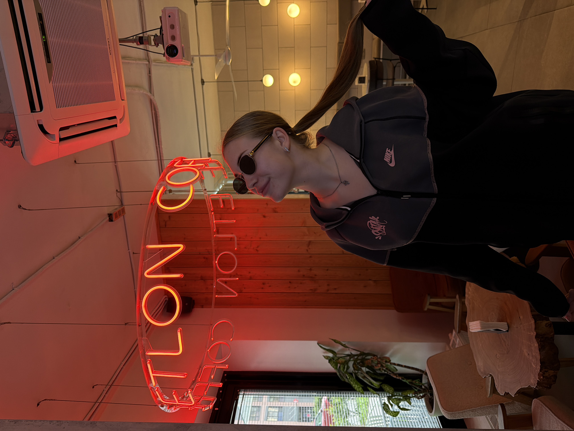 A young woman with blonde hair in a ponytail and sunglasses is standing in a cafe. She is wearing a black and gray Nike hoodie. The cafe has red neon signs that spell out "ETLON COF" and "COF NОЛТЕ". There is a wooden table with a stack of books on it, and several chairs. An air conditioner is mounted on the ceiling, and a projector is hanging from the ceiling.