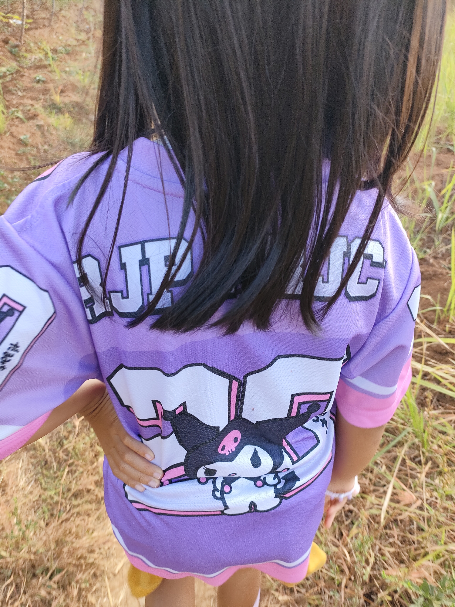 The image shows the back of a young girl wearing a purple t-shirt with a large graphic print. The graphic features a cartoon character with a black head, white face, pink eyes, and a pink skull motif. The t-shirt has white lettering on the back that appears to be part of a larger design, with some letters partially visible. The girl has long, dark hair that covers part of the t-shirt. She is standing outdoors in a grassy, somewhat dry environment, possibly a field or park. The background consists of dry grass and dirt. The lighting suggests it is daytime. The specific location is not discernible.