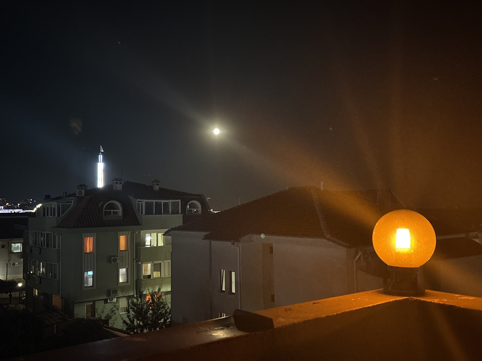 An outdoor nighttime scene captures the facade of several multi-story buildings in Sveti Vlas, Bulgaria, under a dark sky illuminated by a distant moon and a nearby street lamp. The street lamp in the foreground is a spherical, orange-hued fixture with a bright white light emanating from its center. It sits on a textured, reddish-brown ledge or railing.
Behind the lamp, a building with a dark tiled roof and a light-colored facade is visible, featuring several windows, some of which are illuminated. To the left, a taller, pale green building is prominent. It has a more complex architectural design with multiple windows and balconies, some of which emit warm orange and white light, suggesting occupied apartments. Air conditioning units are mounted on the exterior walls. A small tree with sparse foliage stands in front of this building.
Further in the background, a tall, illuminated tower with a flag at its peak pierces the dark sky. The moon is visible as a bright disc in the sky, with rays of light fanning out from it. The overall atmosphere is one of a quiet urban night.