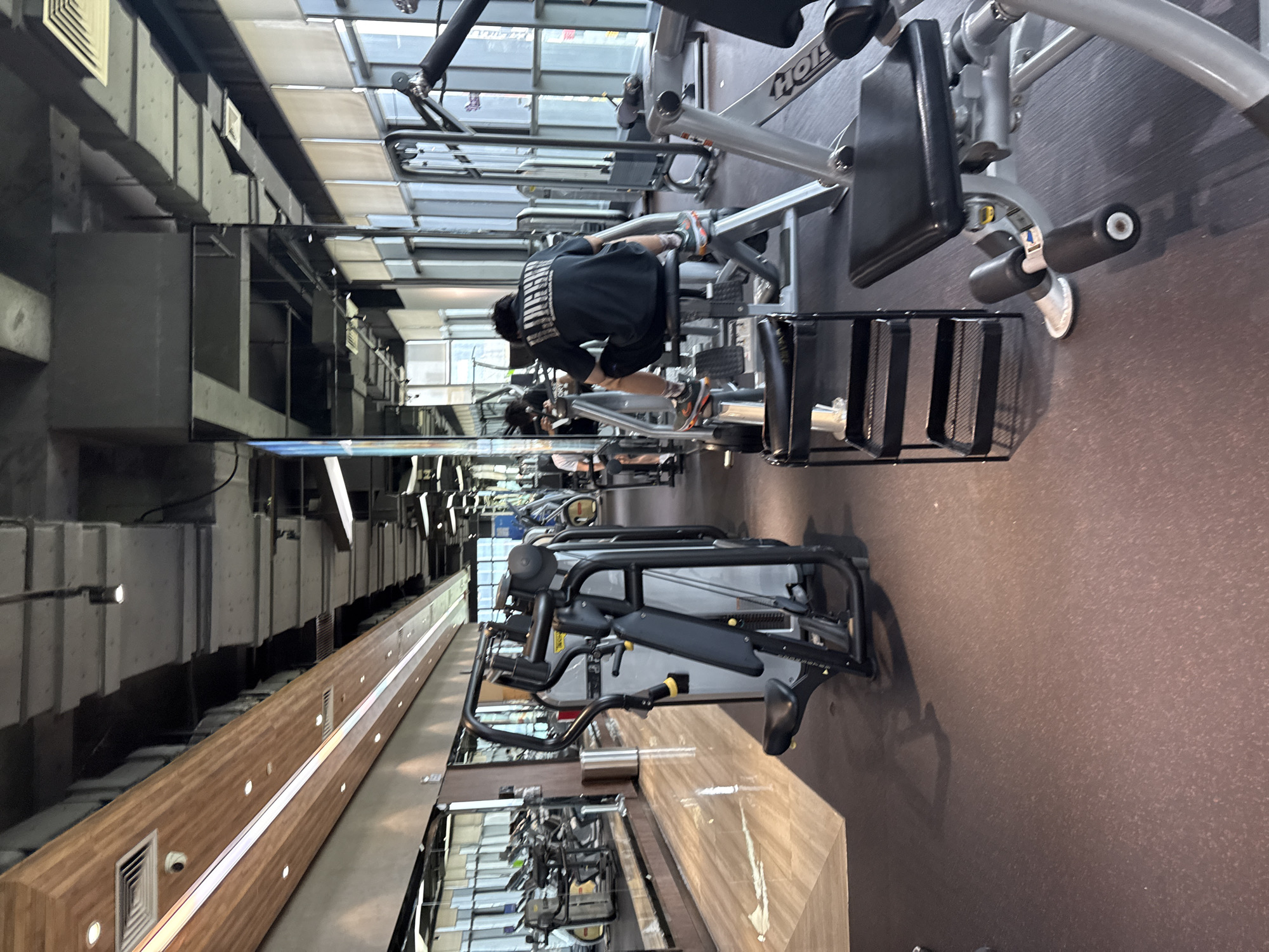 The image shows the interior of a modern gym located in Haizhu District, China. The gym features a variety of weight training machines and cardio equipment, including treadmills and elliptical machines, with a mirrored wall reflecting the space. The floor is covered with a dark, textured material, and the ceiling is industrial with exposed ductwork and lighting fixtures. Natural light streams in from large windows along the side of the building. In the foreground, a person wearing a black t-shirt and shorts is seated on a weight machine, facing away from the camera. Other gym-goers and equipment are visible in the background, creating a bustling, active atmosphere.