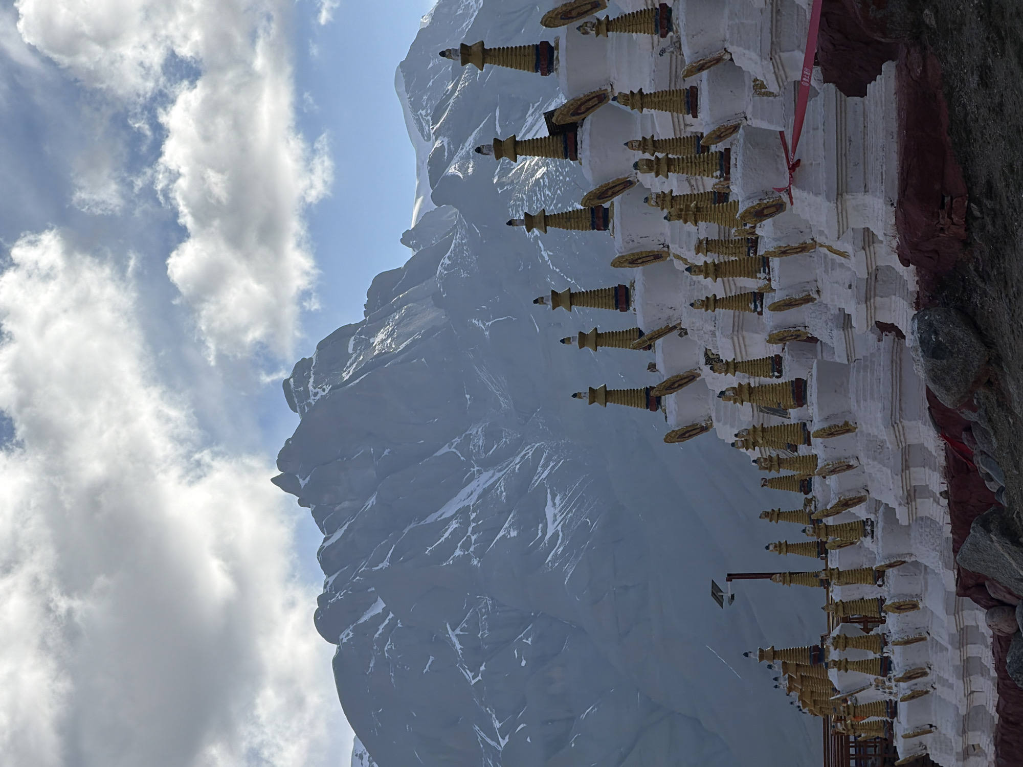 The image shows a snowy mountain peak under a partly cloudy sky adjacent to a white building with several golden spire-shaped structures atop it. The building is situated on a rocky area. The picture is rotated approximately 90 degrees counter-clockwise. There is no clear evidence to confirm this image is related to Parga town in China.