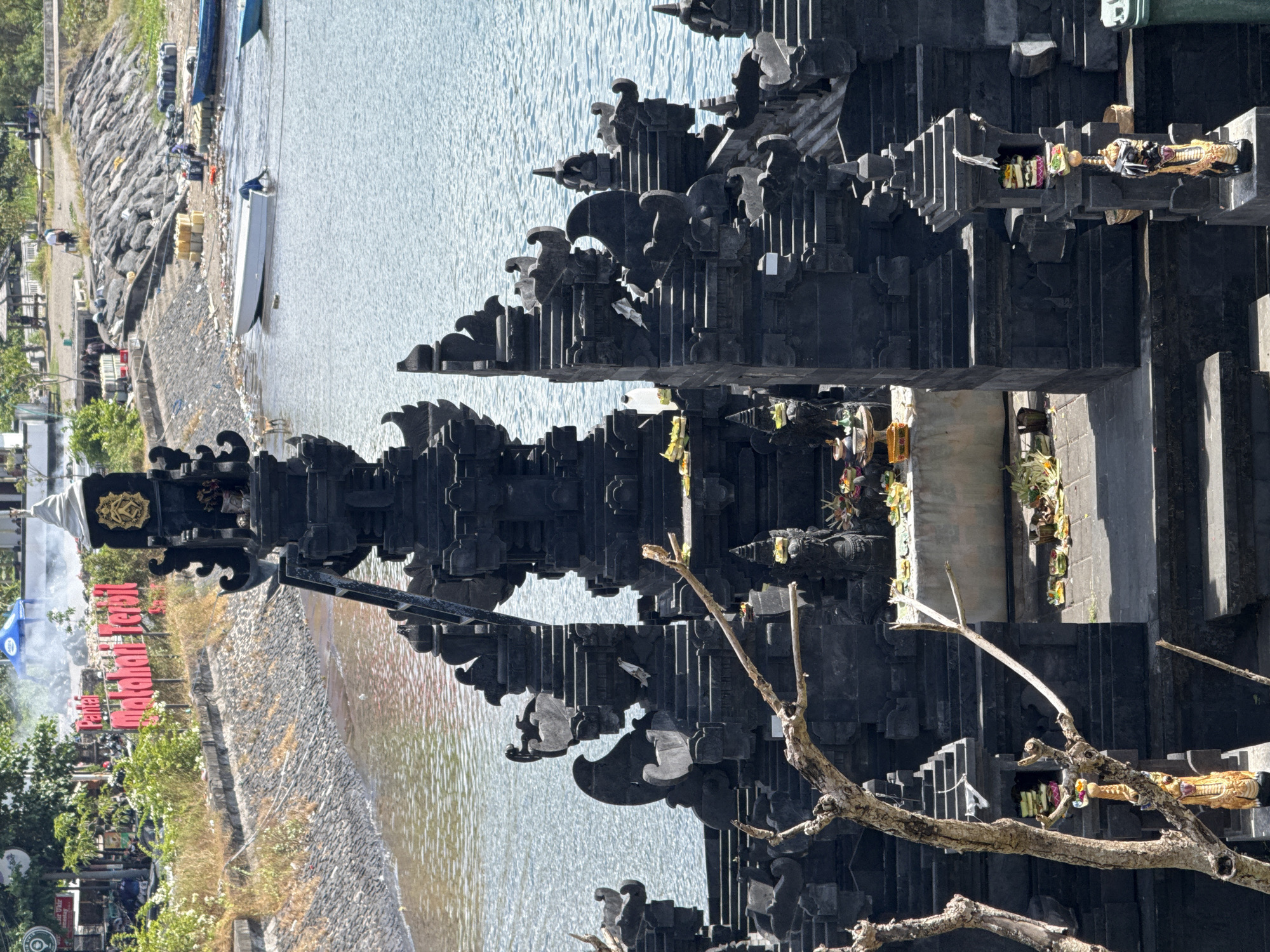 This photograph captures a structure near the water at Pantai Matahari Terbit, in Denpasar, Indonesia. The structure is built in a series of stacked, carved levels, appearing dark in color. Along the side of the structure are religious figures, and offerings of flowers and food. In the background, the coastline is visible with a boat docked near a stony embankment. Along the shore, signage and small shops are visible, suggesting a tourist or recreational area. The water appears calm and light blue.