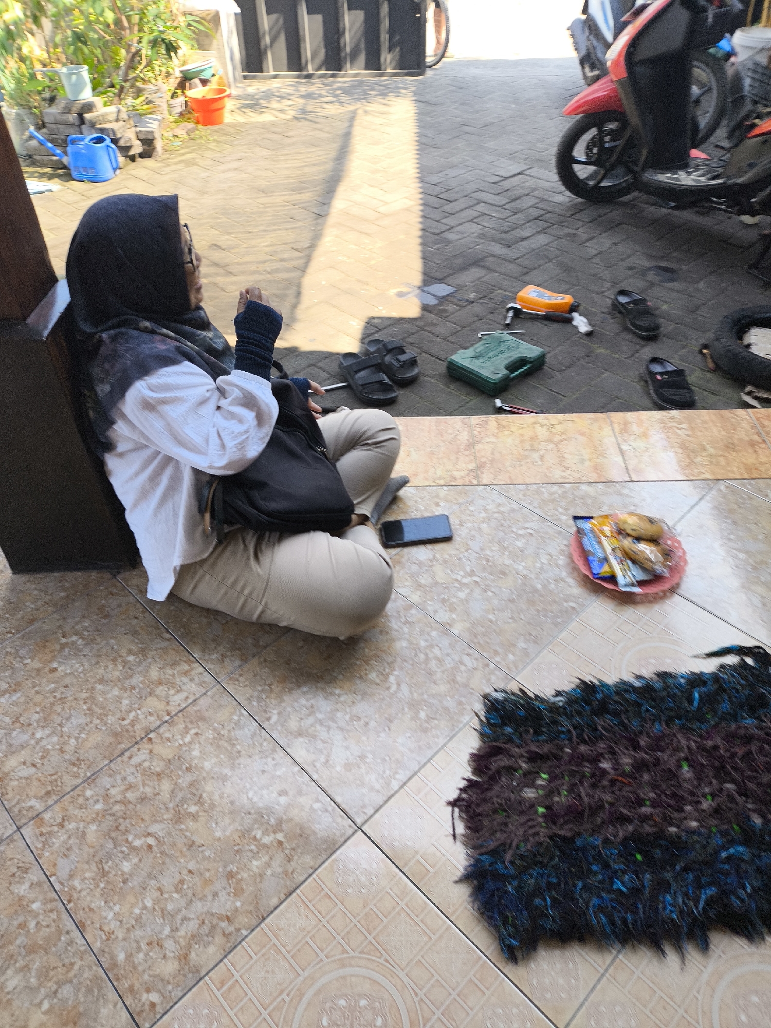 In Lumajang, Indonesia, a woman wearing a hijab and glasses sits cross-legged on a tiled floor next to a dark wooden pillar. She is dressed in a white long-sleeved shirt and beige pants, with a black bag resting on her lap. Her right hand is raised and her fingers are slightly curled, while her left hand is obscured by the bag. In front of her, on the tiled ground, is a black smartphone. Further away, on a paved area with brick patterns, several objects are scattered: a green toolbox, an orange object resembling a tool or device, a black flip-flop, a pair of black sandals, and a red circular plate holding bananas and packaged snacks. A red and black scooter is parked in the background, with its front wheel visible. To the left of the woman, a blue watering can and several potted plants are visible on a stack of bricks. The environment appears to be an outdoor area, possibly a yard or a terrace, with sunlight casting shadows.