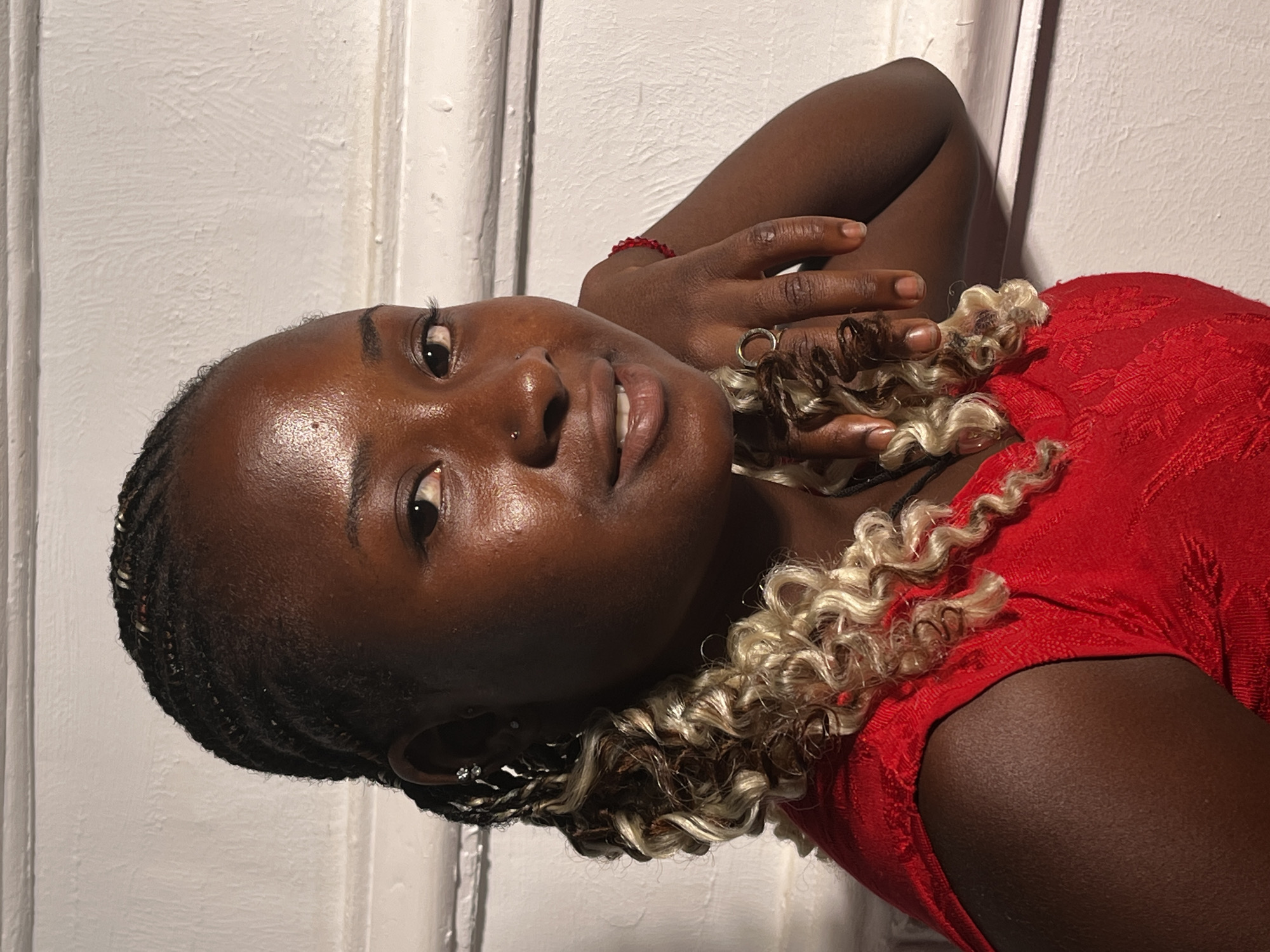 A dark-skinned woman lies on her side against a white wall, possibly indoors. She is wearing a red patterned top. Her hair is styled in braids with blonde curly extensions. She has a nose piercing, and she's wearing a ring. She has one hand near her head and is wearing a red bracelet. The lighting is somewhat dim. The location in the city of Barakin Akawo, Nigeria, cannot be verified from the image.