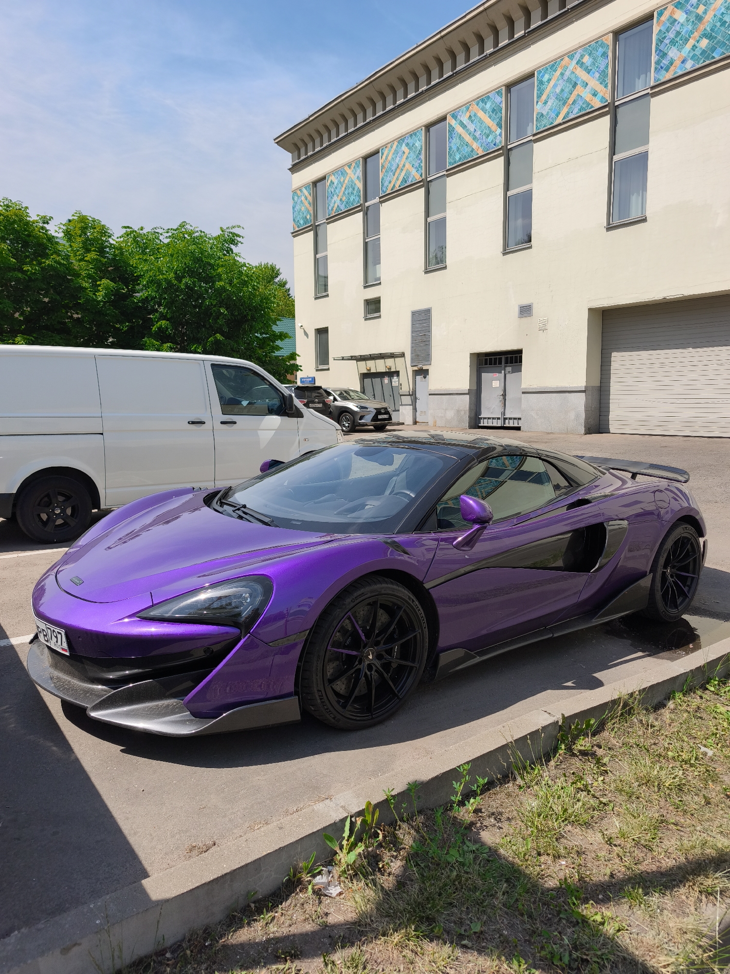 A brightly lit outdoor scene depicts a metallic purple McLaren sports car parked on an asphalt surface next to a concrete curb. The McLaren features black multi-spoke wheels, a black roof, carbon fiber accents including a front splitter, side skirts, and a rear spoiler. A Russian license plate, "PB797," is visible on the front.
Behind the McLaren, a white passenger van, possibly a Volkswagen Transporter, is parked. Further in the background, a light grey SUV or crossover is partially visible.
The background is dominated by a multi-story building with light cream-colored walls and a decorative cornice along the roofline. The facade features several rectangular windows, above which are panels adorned with a geometric pattern in shades of turquoise and orange/tan. The ground level of the building includes a large light grey roll-up garage door, a darker access door, and various vents.
To the left of the building and behind the white van, lush green deciduous trees are visible against a light blue sky with scattered wispy white clouds. The asphalt parking area is separated by a concrete curb from a narrow strip of dry grass and weeds. Distinct shadows are cast across the asphalt and curb, indicating bright sunlight. No individuals are present in the scene. The location is in the city of Unknown, Unknown.