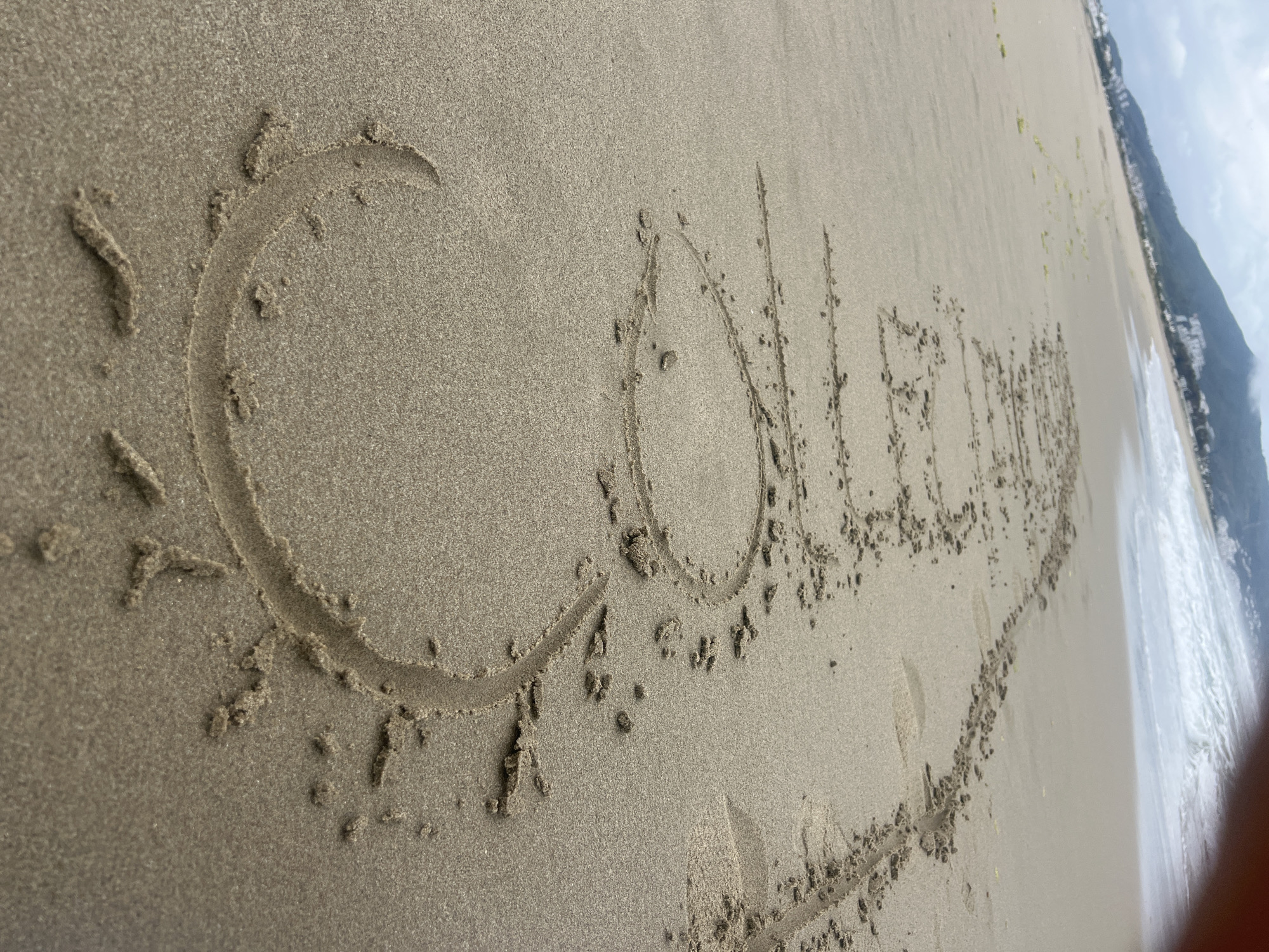 A close-up, oblique shot captures a word and a symbol meticulously drawn into the damp, light brown sand of a beach. The main subject is the word "Collegium" written in capital letters, extending across the frame from left to right. To the immediate left of the initial capital 'C', a stylized sun symbol with a circular center and radiating lines is also etched into the sand. The grooves of the writing and drawing are distinct, showing the texture of the displaced sand.
The setting is a sandy beach, likely at the edge of the water, indicated by the dampness of the sand. In the mid-ground, a calm body of water, presumably the sea, is visible with gentle waves breaking onto the shore. Further in the background, a distant coastline characterized by mountains or hills stretches across the horizon under a bright, possibly partly cloudy, sky. The lighting suggests it is daytime. The scene conveys a relaxed beach atmosphere. No individuals are visible in the image. The location context indicates this is likely Martil, Morocco.