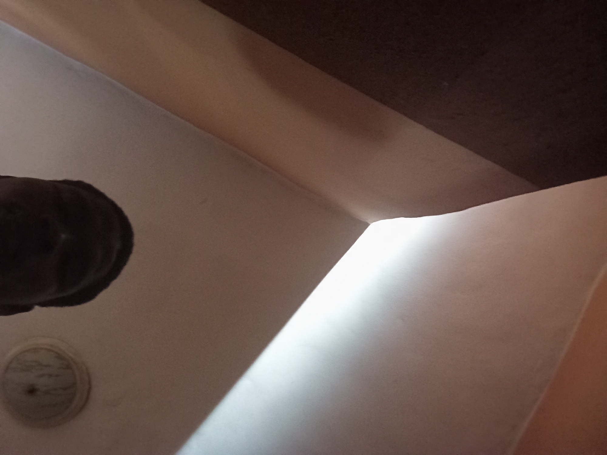 The image presents an abstract, upward-angled view within an interior architectural space, likely focusing on a ceiling and upper wall corner. The scene is characterized by intersecting planes and contrasting tones of dark brown, lighter brown/beige, and white.
In the upper section, a dark brown, textured surface, possibly a ceiling or a prominent wooden beam, stretches across the frame. Adjacent to it, a lighter brown or beige surface forms an angular plane, suggesting an adjoining wall or another structural element. These darker elements delineate the upper part of the visible interior.
A significant feature is a strong, bright shaft of light that diagonally cuts across the right side of the frame, illuminating a white or off-white wall or ceiling surface. This intense illumination suggests natural light, likely sunlight, entering the room during daytime, creating a dramatic contrast with the shadowed areas.
Two discernible objects are present in the lower-left portion of the image. A dark, indistinct, and somewhat globular object appears to hang or protrude from the upper-left, possibly a light fixture or a component of a ceiling fan. Below it, a circular, light-colored object with a faint patterned surface is mounted on the lighter wall or ceiling, resembling a recessed light fixture cover, a small vent, or a smoke detector.
No people are visible, and no specific activity or event is taking place. The image is a static portrayal of an interior, highlighting architectural details and the effect of light, consistent with an indoor setting in Nkpor, Nigeria. There is no visible text.