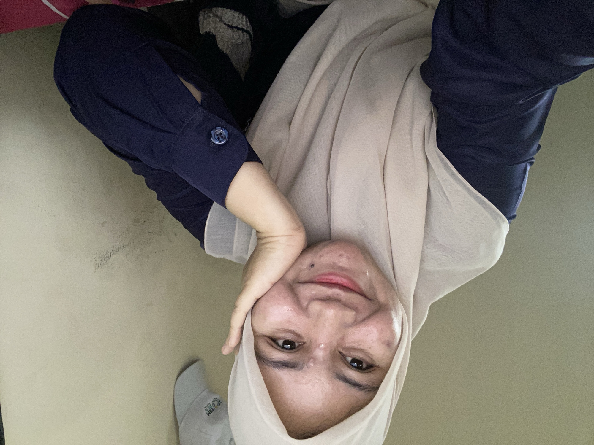 A young woman wearing a beige hijab and a navy blue shirt is photographed from the chest up, in what appears to be an indoor setting. She is smiling and resting her left cheek on her hand, with her eyes looking towards the camera. The background consists of a plain, textured wall, and a light-colored object, possibly a cap, is visible hanging on it to the right. The lighting is soft, casting a warm glow on her face. The image is a selfie, suggesting a personal and informal moment. Based on the metadata, the location is Banjarbaru, Indonesia.